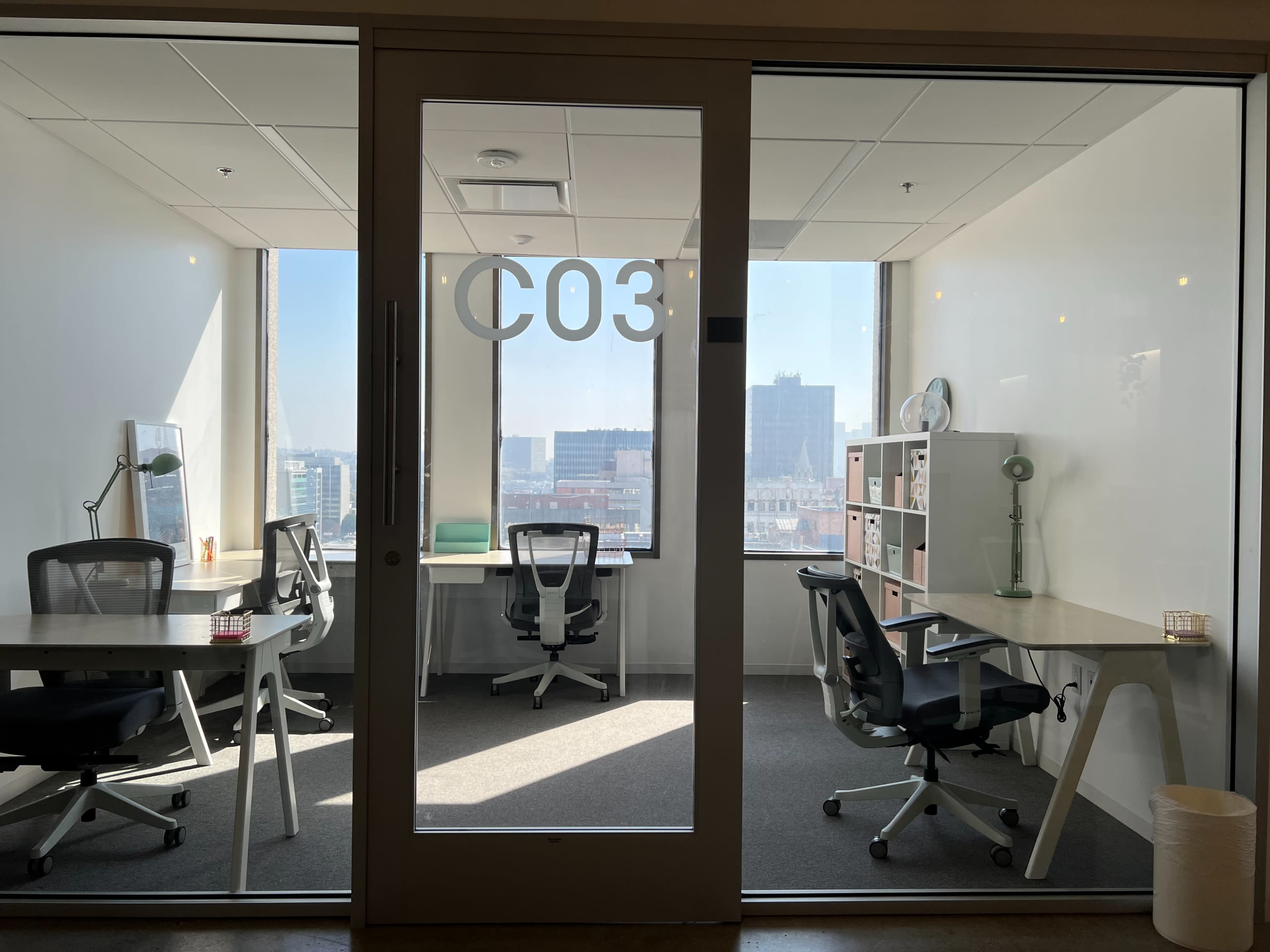 A glass-walled office space labeled "C03" with two desks, each equipped with a chair and a view of the city skyline outside.