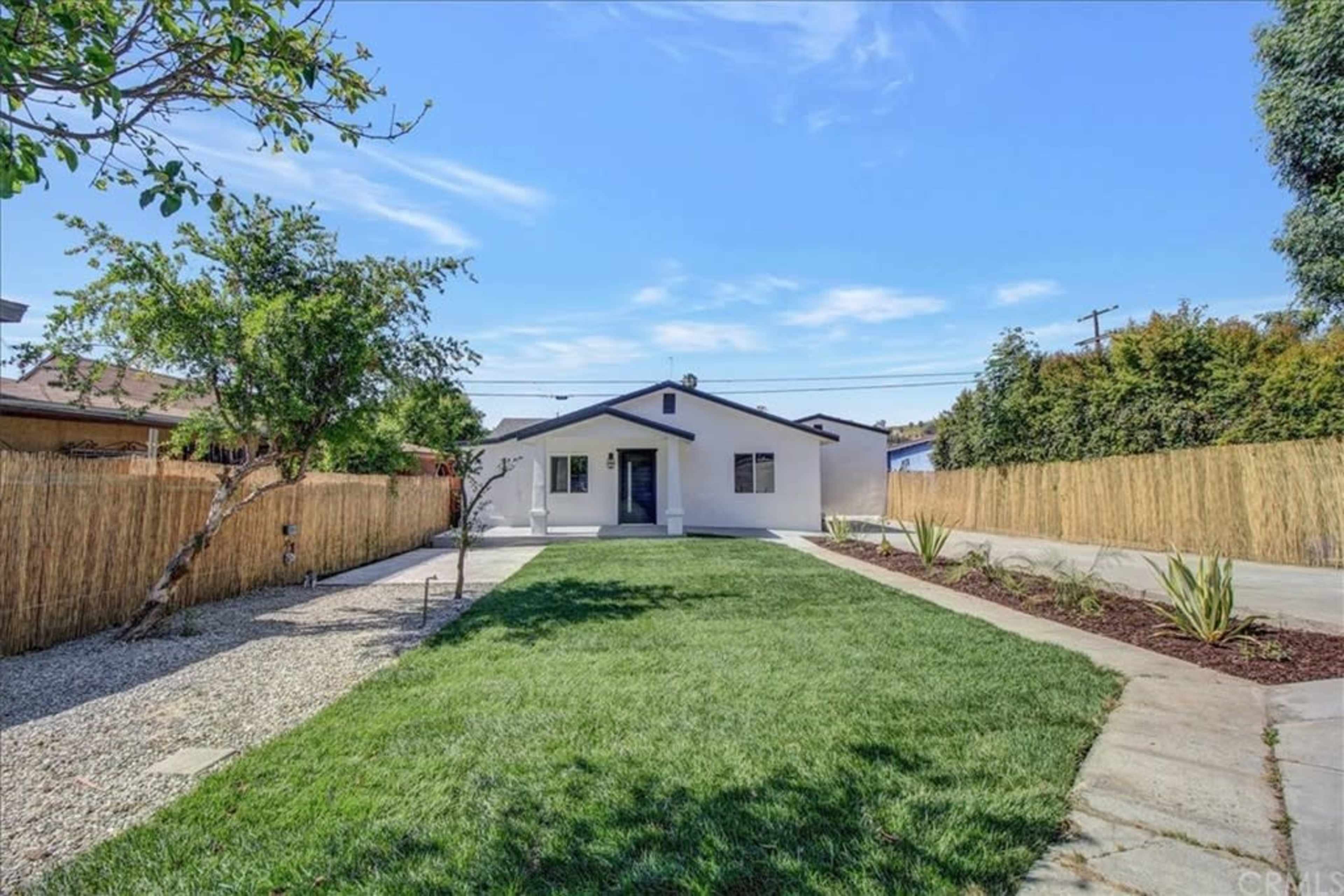The image shows a single-story house with a green lawn surrounded by pathways and landscaped areas, set against a clear blue sky.