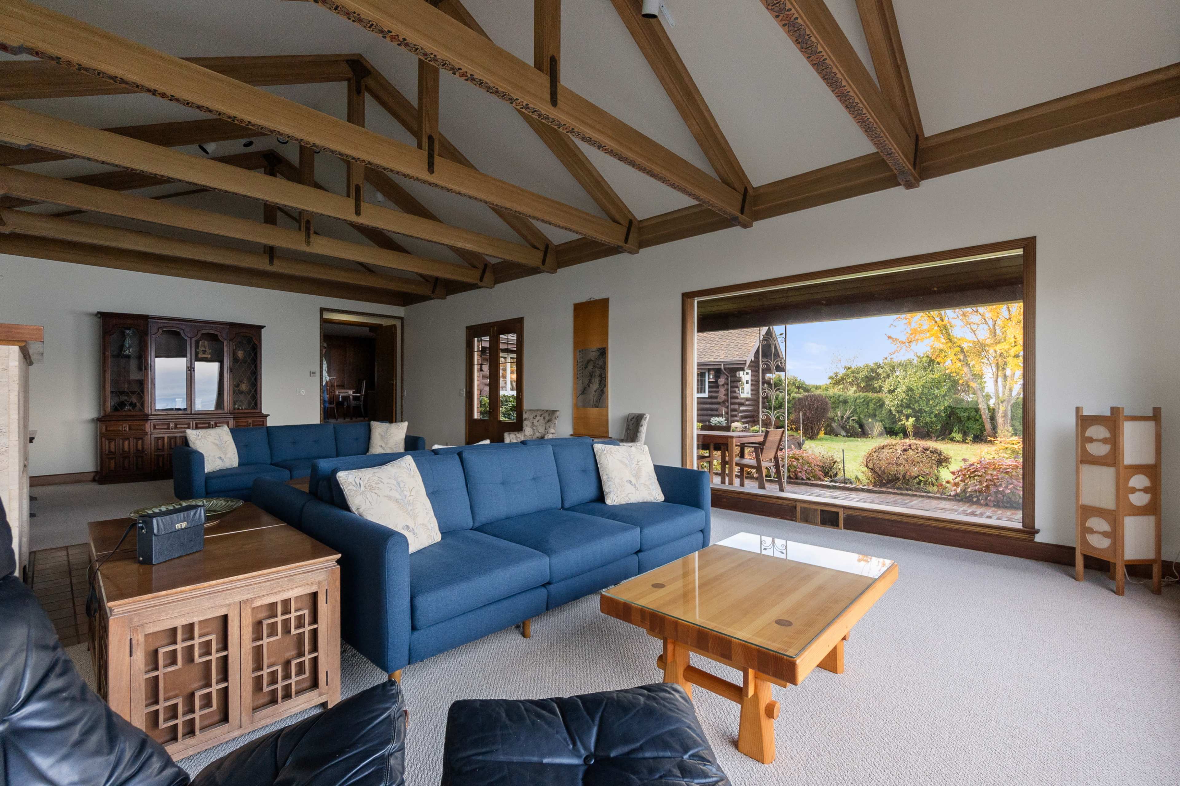 The image shows a spacious living room with a blue sectional sofa, wooden furniture, and large windows that open to a view of a garden.