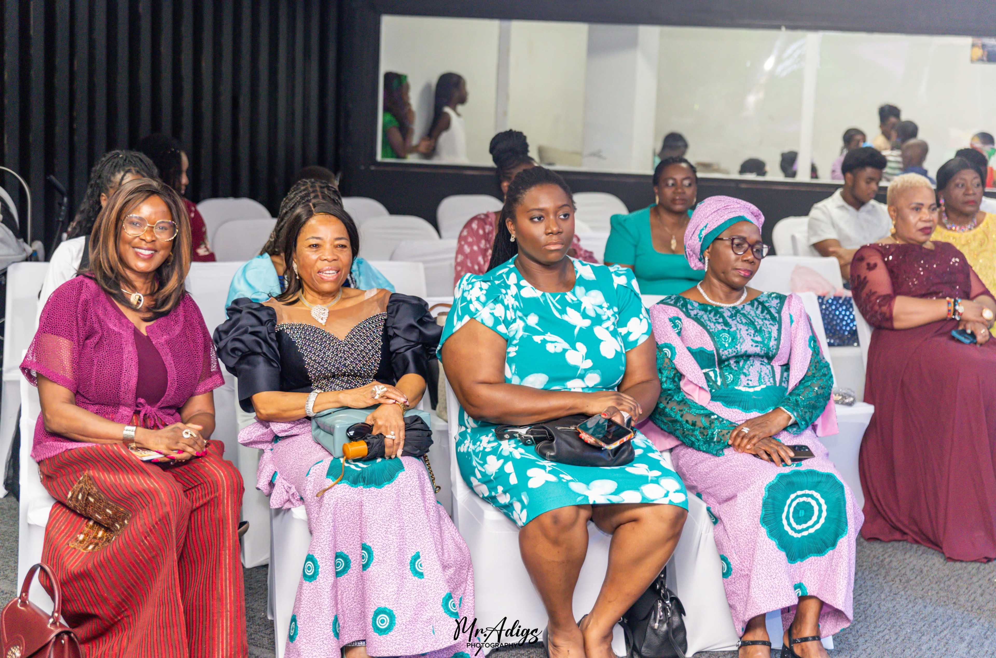 A group of women and attendees are seated in a venue, with some engaged in conversation and others looking toward the front.