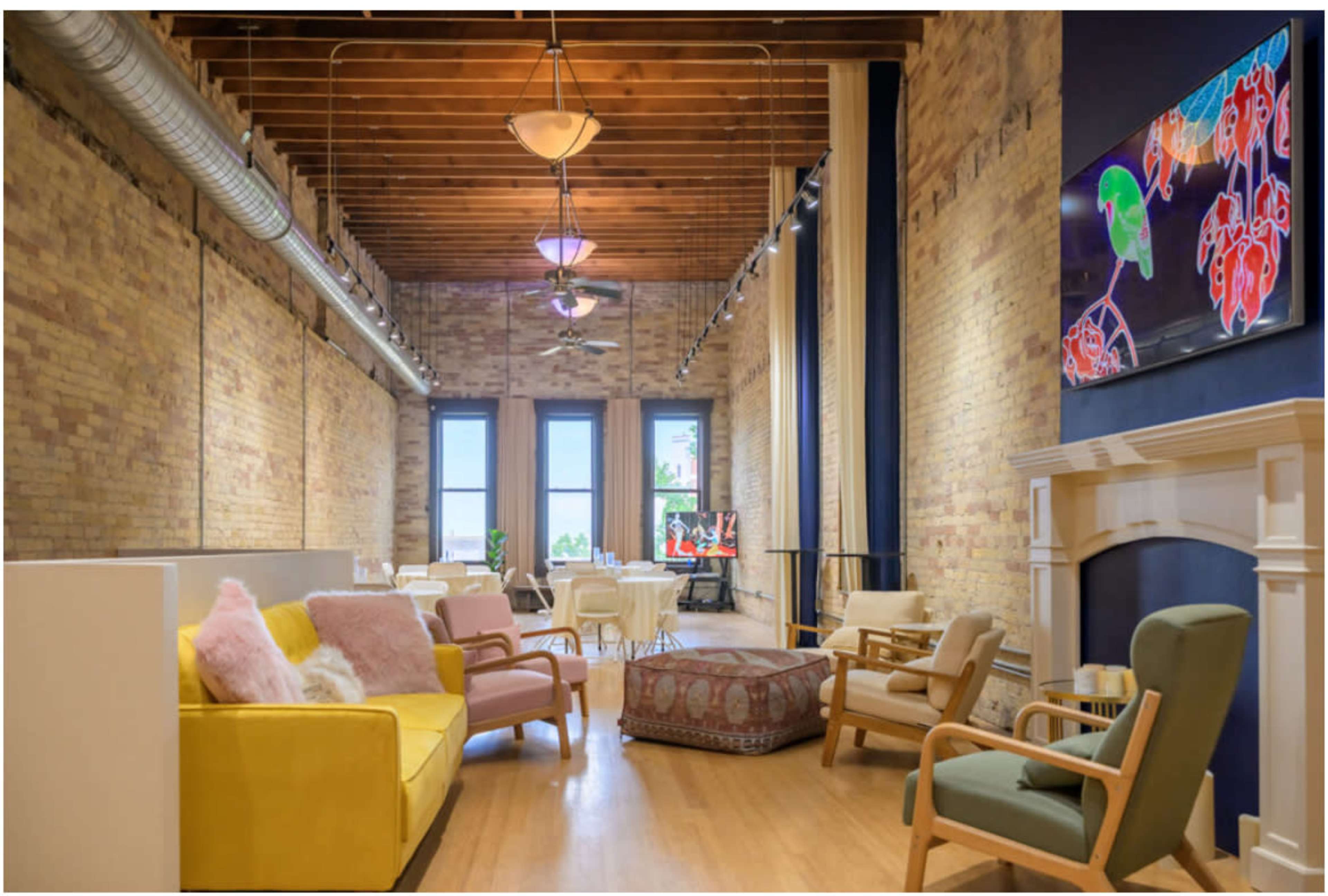 The image shows a spacious, modern lounge area with high ceilings, exposed brick walls, and a variety of seating options, including colorful sofas and a decorative pouf, alongside dining tables and a large television displaying vibrant artwork.