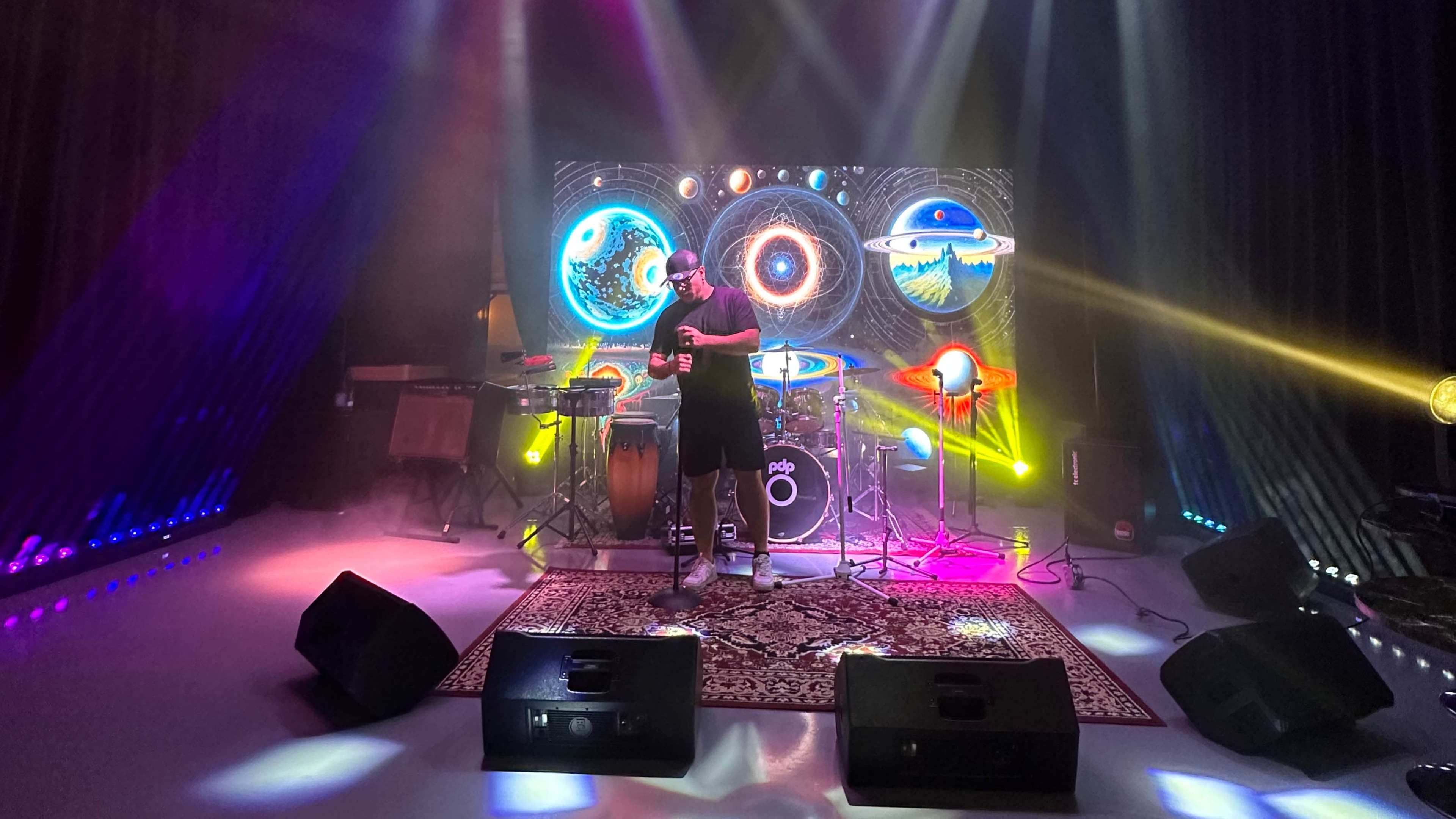 A musician performs on stage in front of a colorful cosmic backdrop, accompanied by various instruments and stage lights.