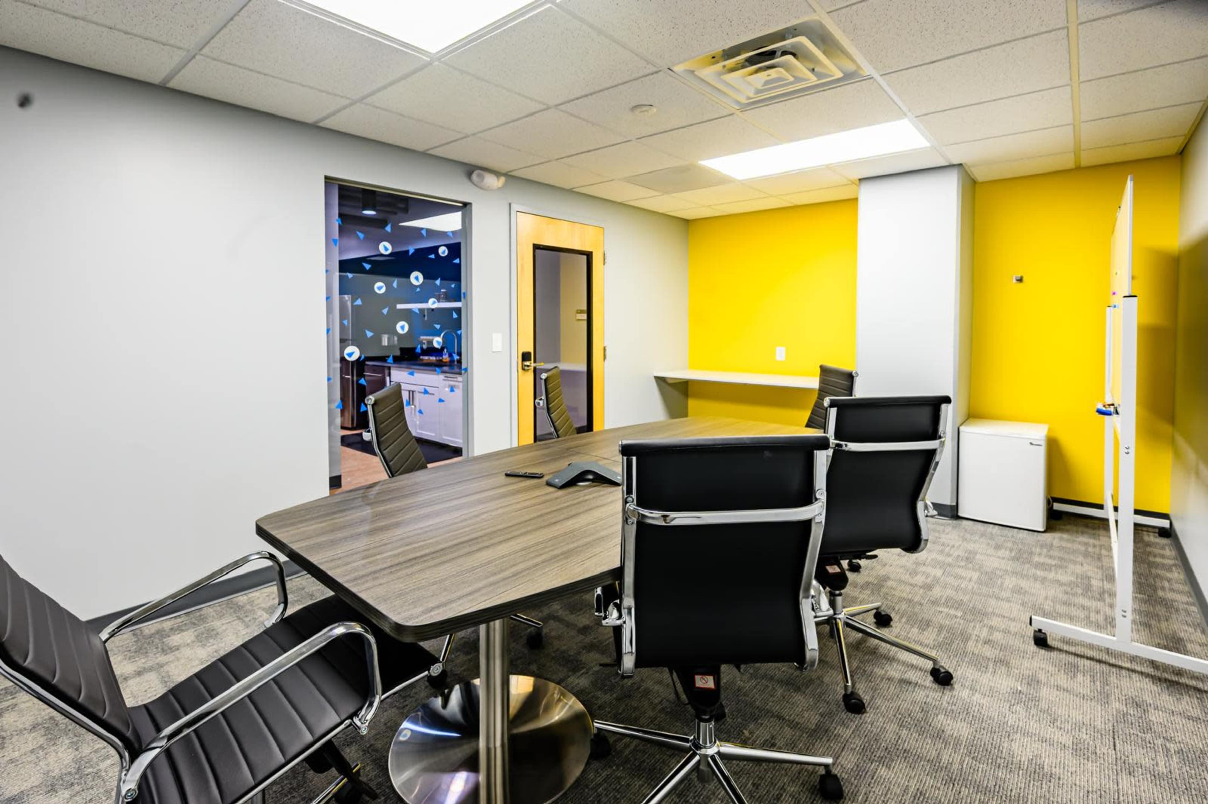 A modern conference room features a large wooden table surrounded by black office chairs, with a yellow wall and a door leading to another room.