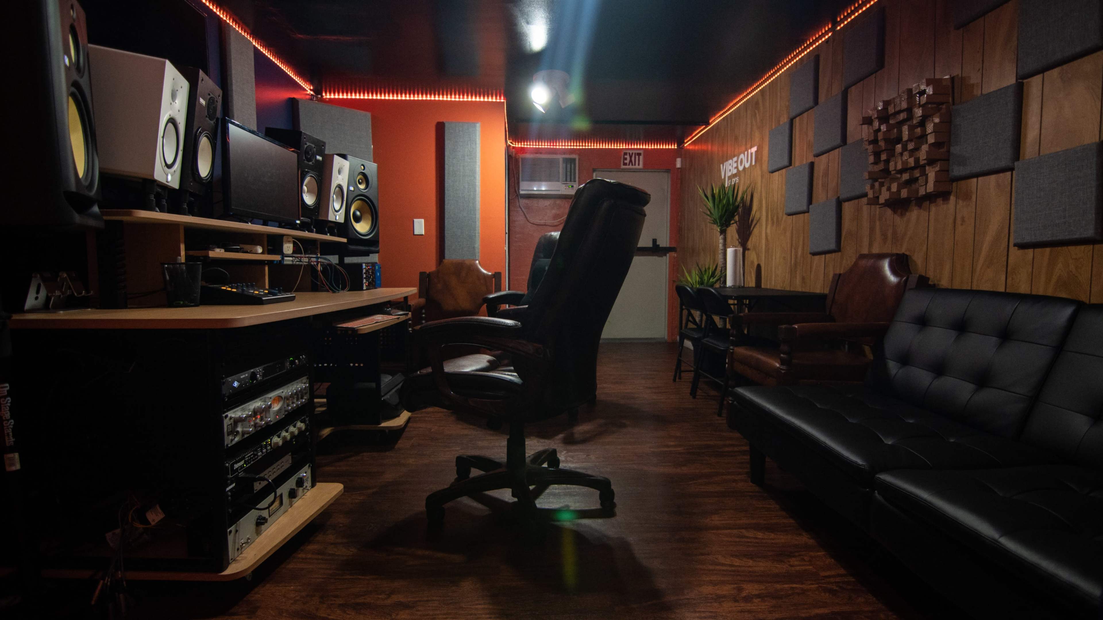 The image shows a music studio with various audio equipment, a comfortable seating area, and warm wooden paneling.