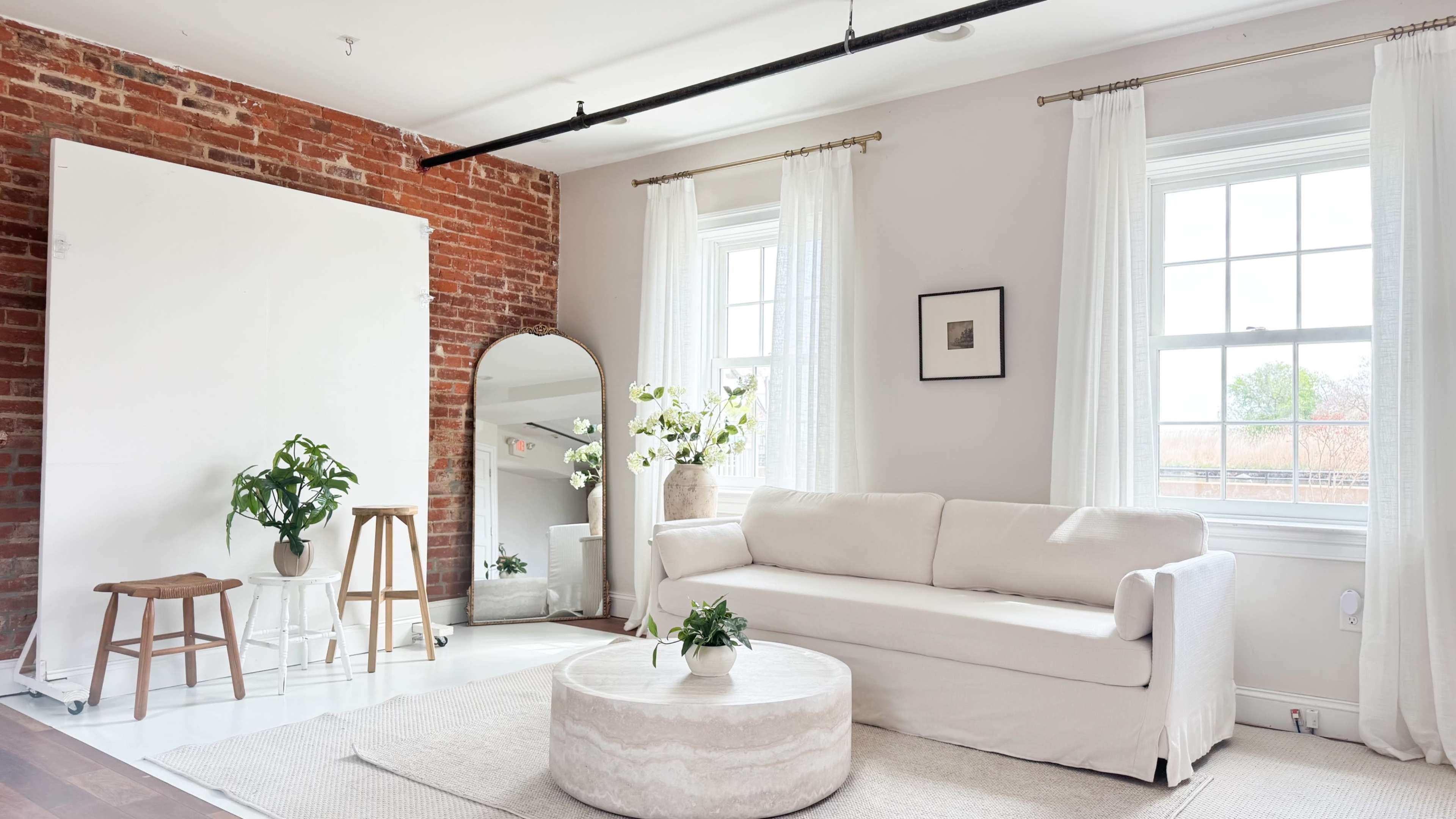 The image shows a bright and airy living room with a white sofa, a round coffee table, two wooden stools, a large mirror, and a vase of flowers, all set against a backdrop of exposed brick walls and large windows.