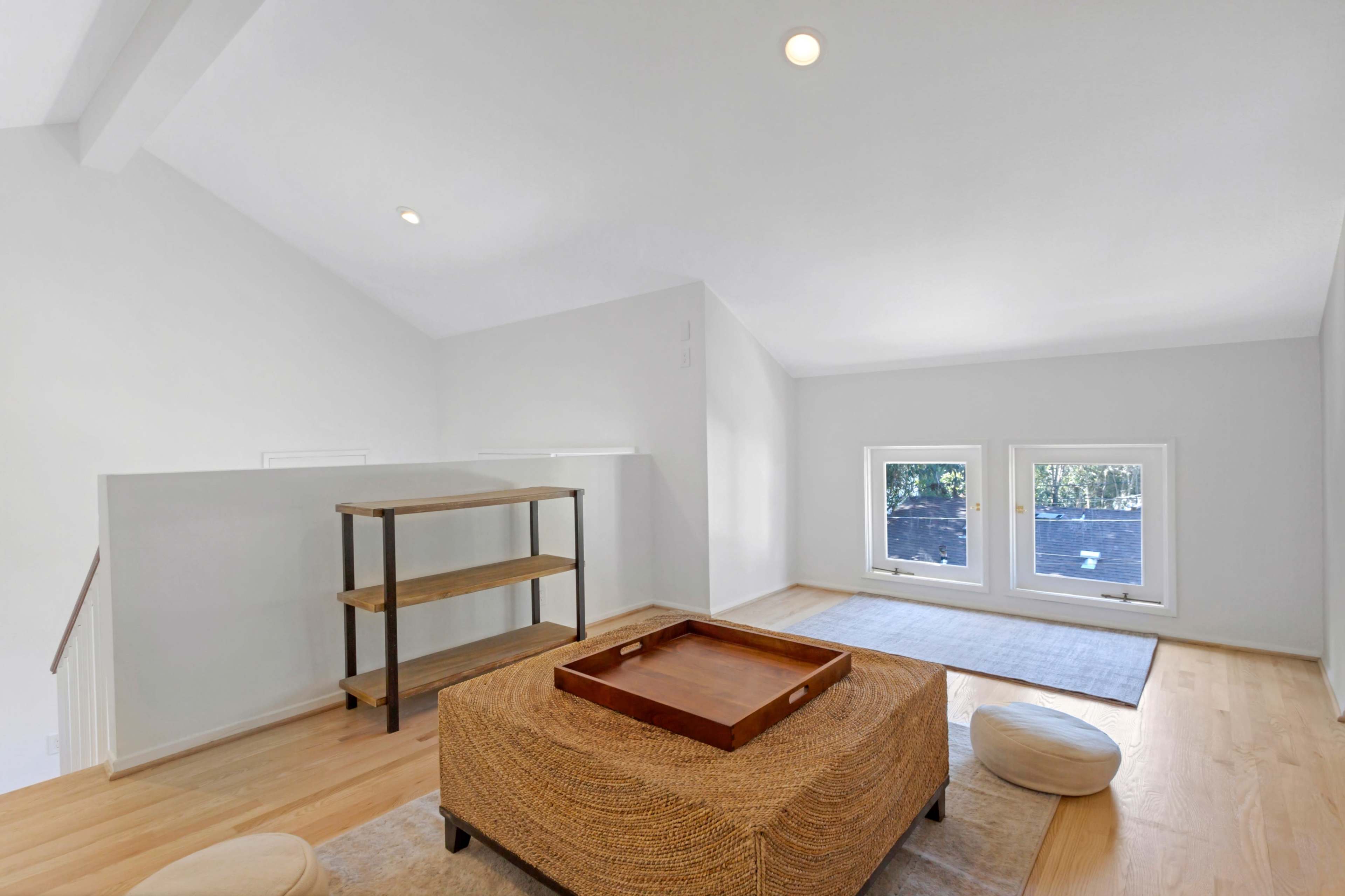 The image shows a bright, minimalist room featuring a wooden table, a woven ottoman, and large windows allowing natural light to enter.