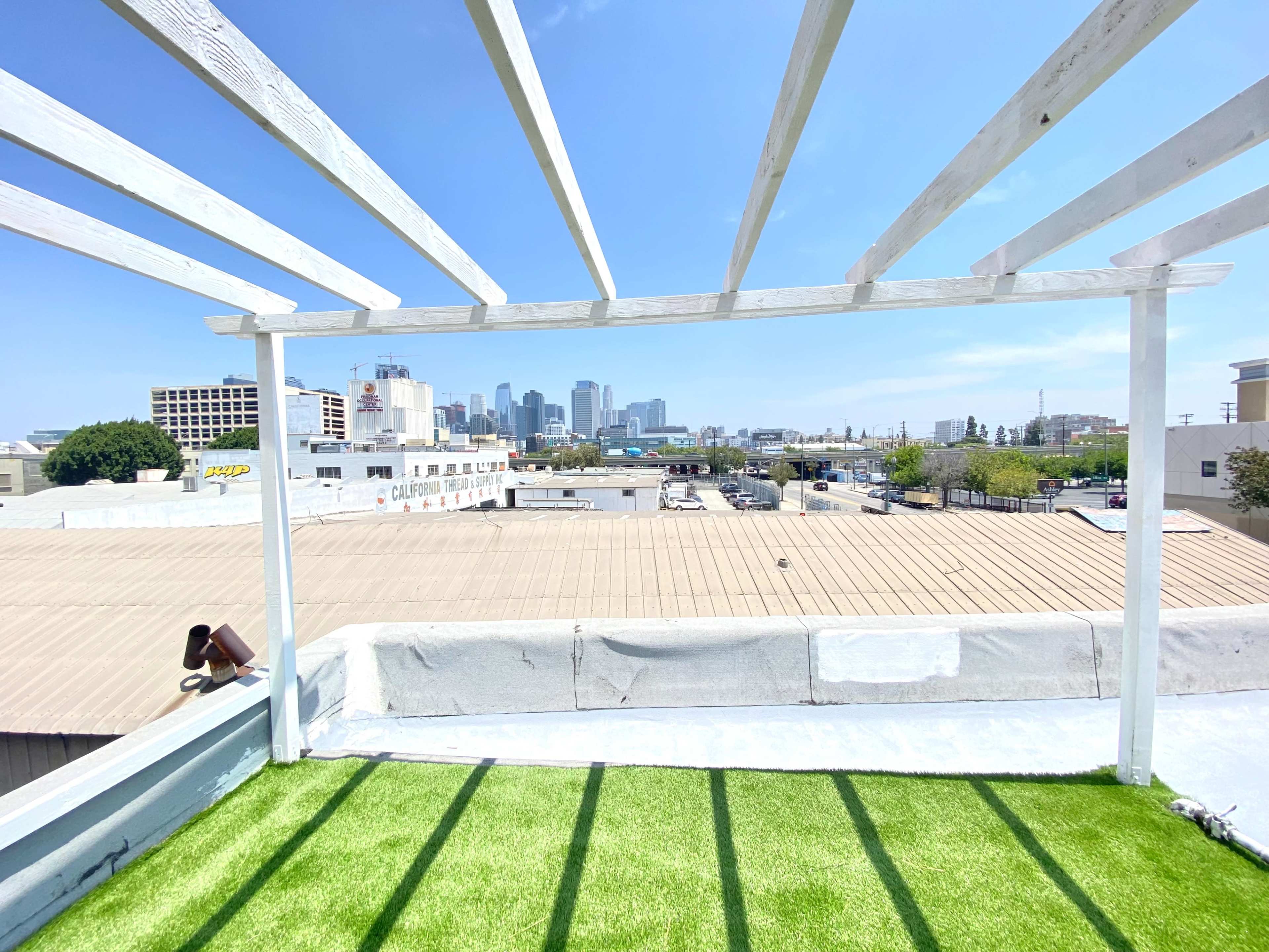 A rooftop terrace with artificial grass, white wooden beams, and a view of a city skyline in the background.