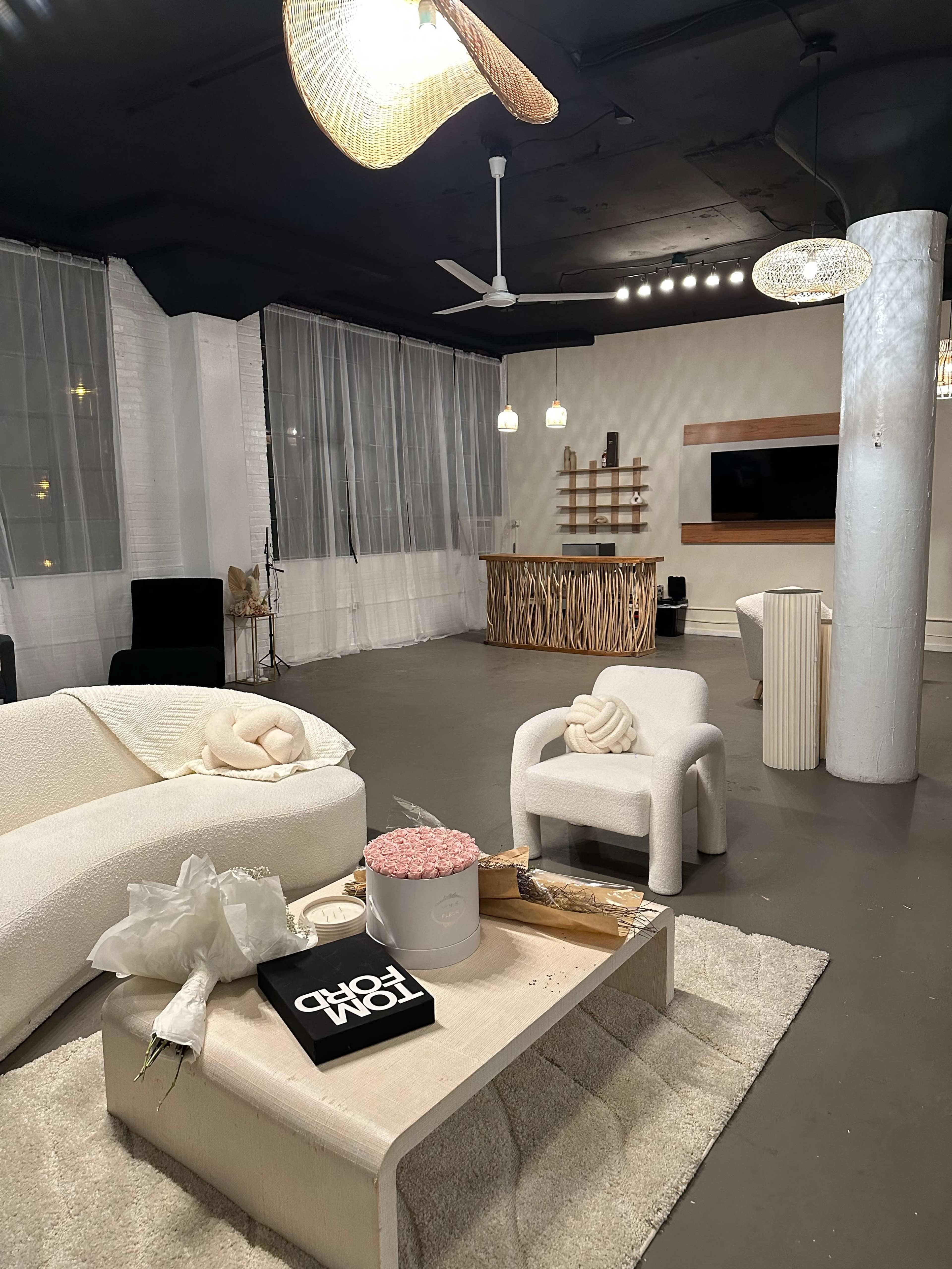 The image shows a modern, minimalist lounge area featuring a light-colored sofa, a coffee table with decorative items, and a wooden bar in the background, all under soft lighting.