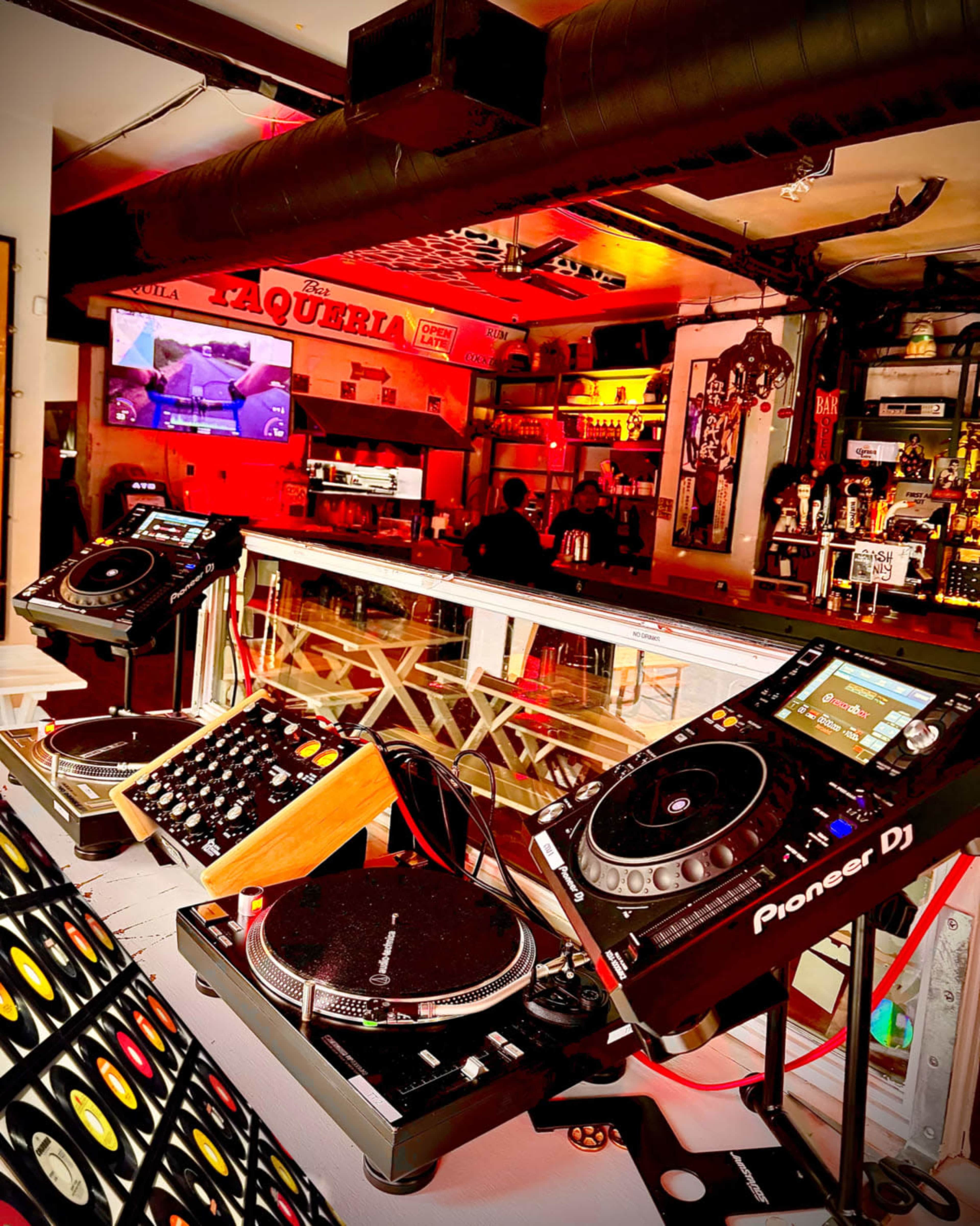 The image shows a DJ setup with turntables and a mixer, positioned in a vibrant bar that features colorful lighting and a visible display screen in the background.