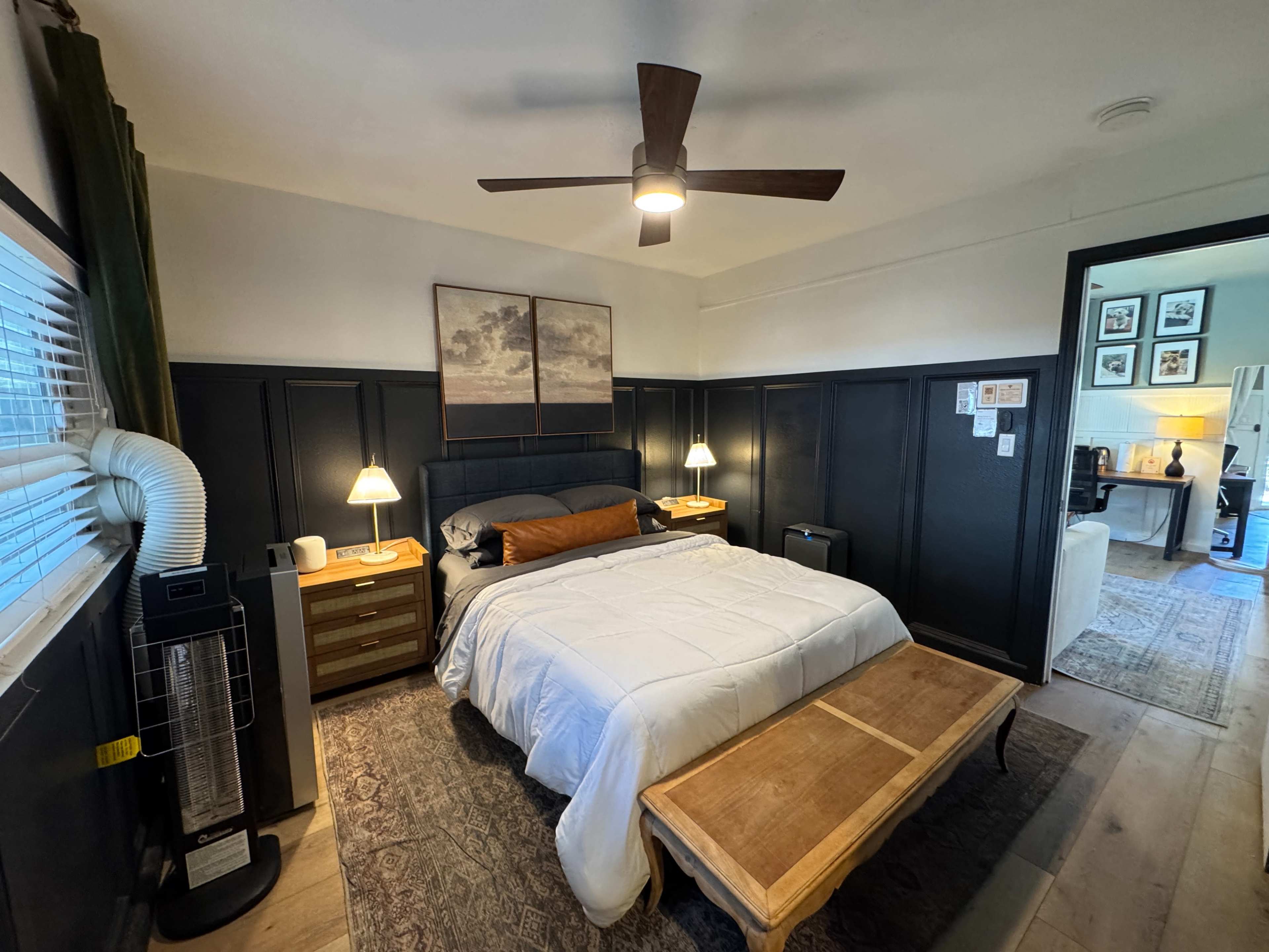 The image shows a neatly arranged bedroom with a bed, nightstands, a coffee table, and a workspace visible in the background.
