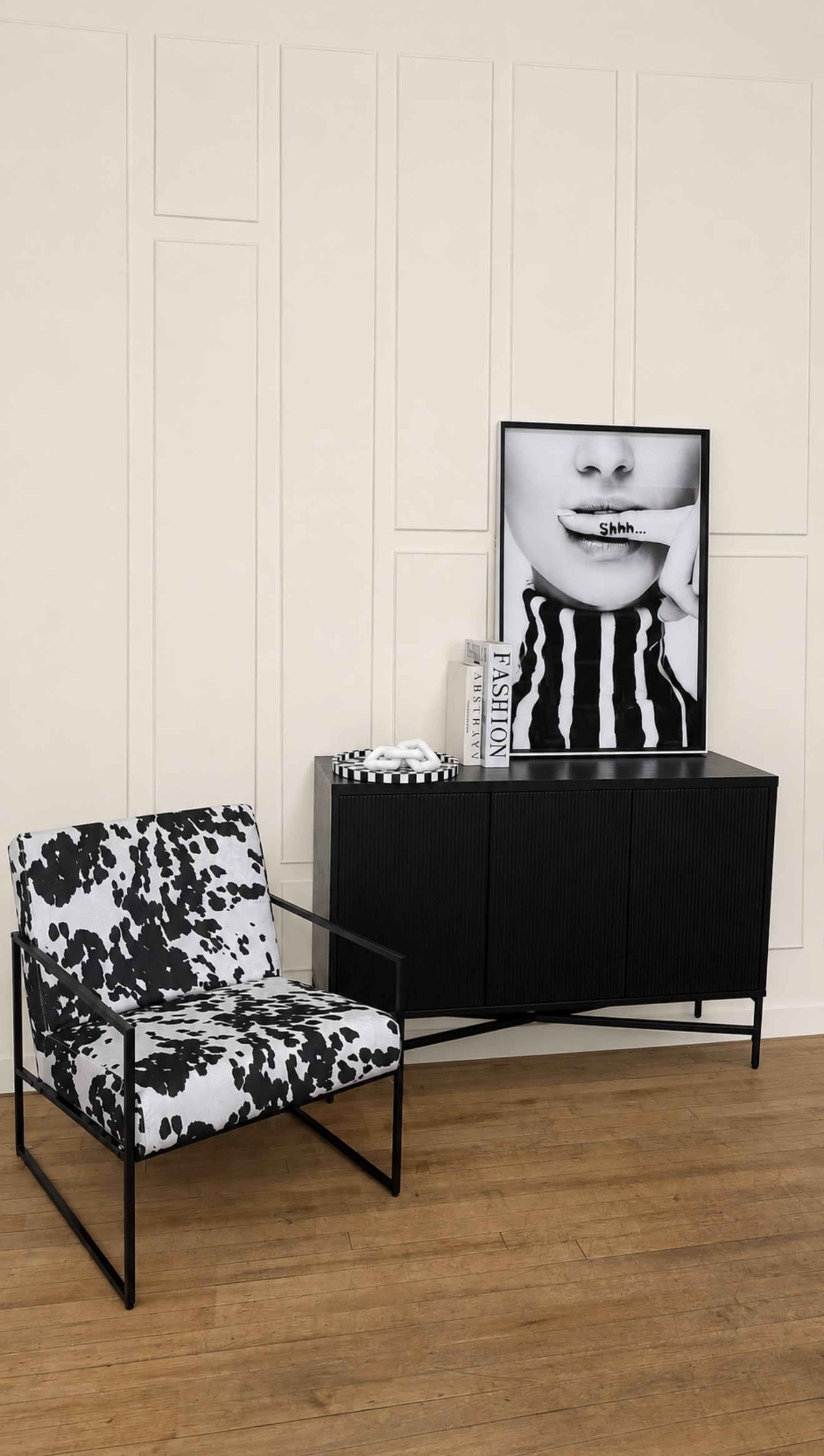 The image features a black and white patterned chair next to a sleek black cabinet, with a framed black and white artwork of a face on the wall above.