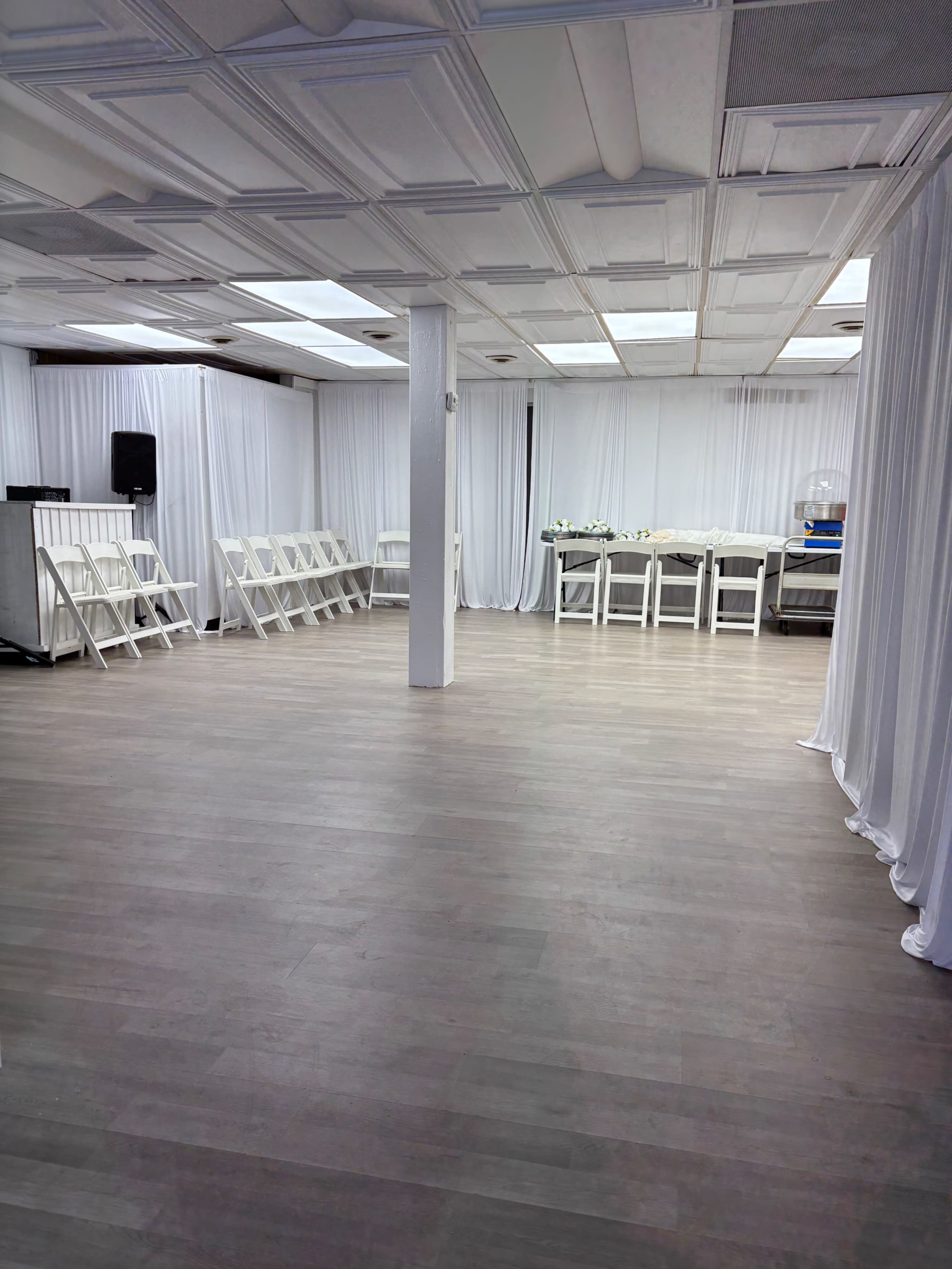 The image shows an empty event space with white drapery and wooden flooring, featuring a row of folding chairs on one side and a food setup area on the opposite side.