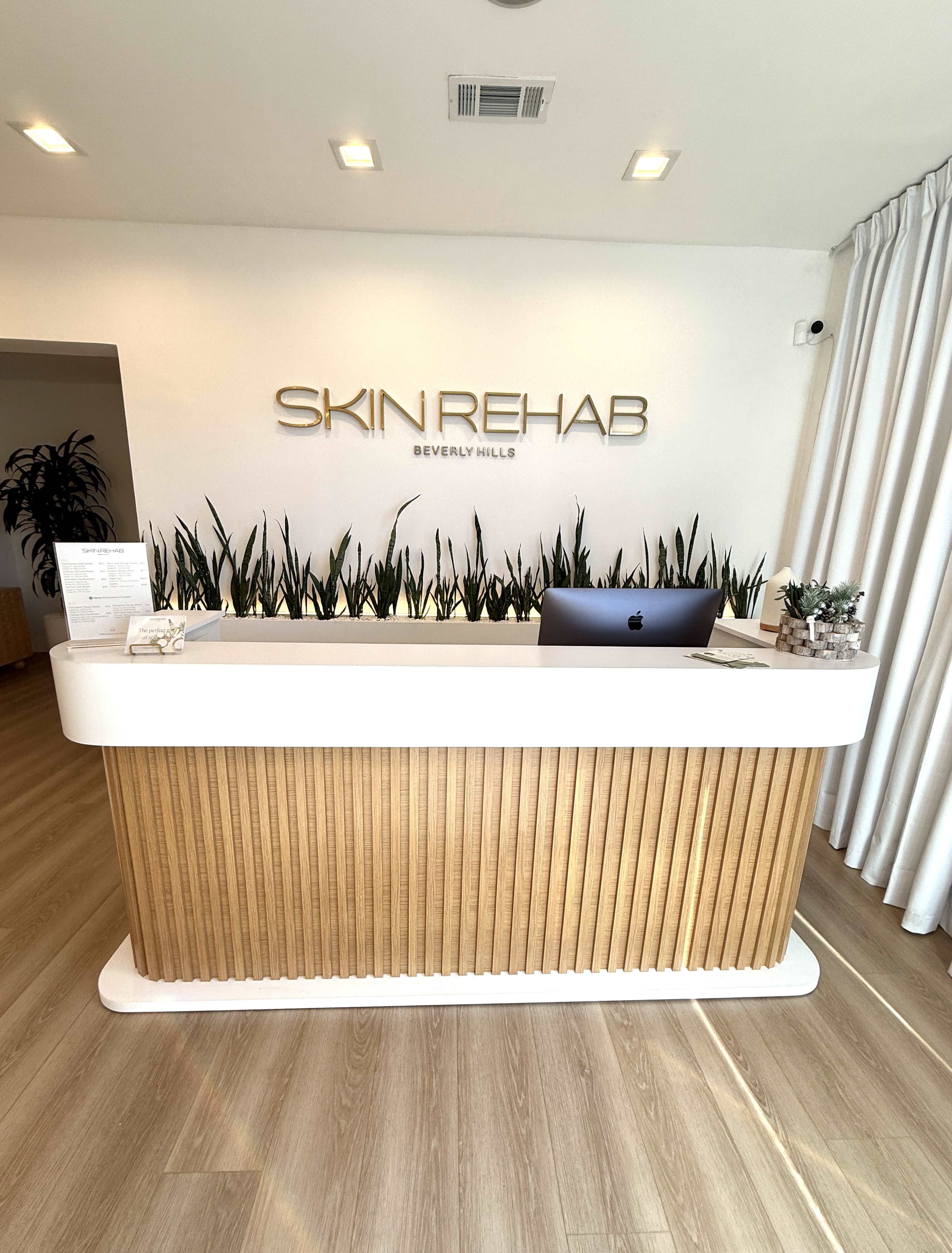 The image shows a modern reception desk at Skin Rehab in Beverly Hills, featuring a sleek design with wooden accents and a computer on the countertop.