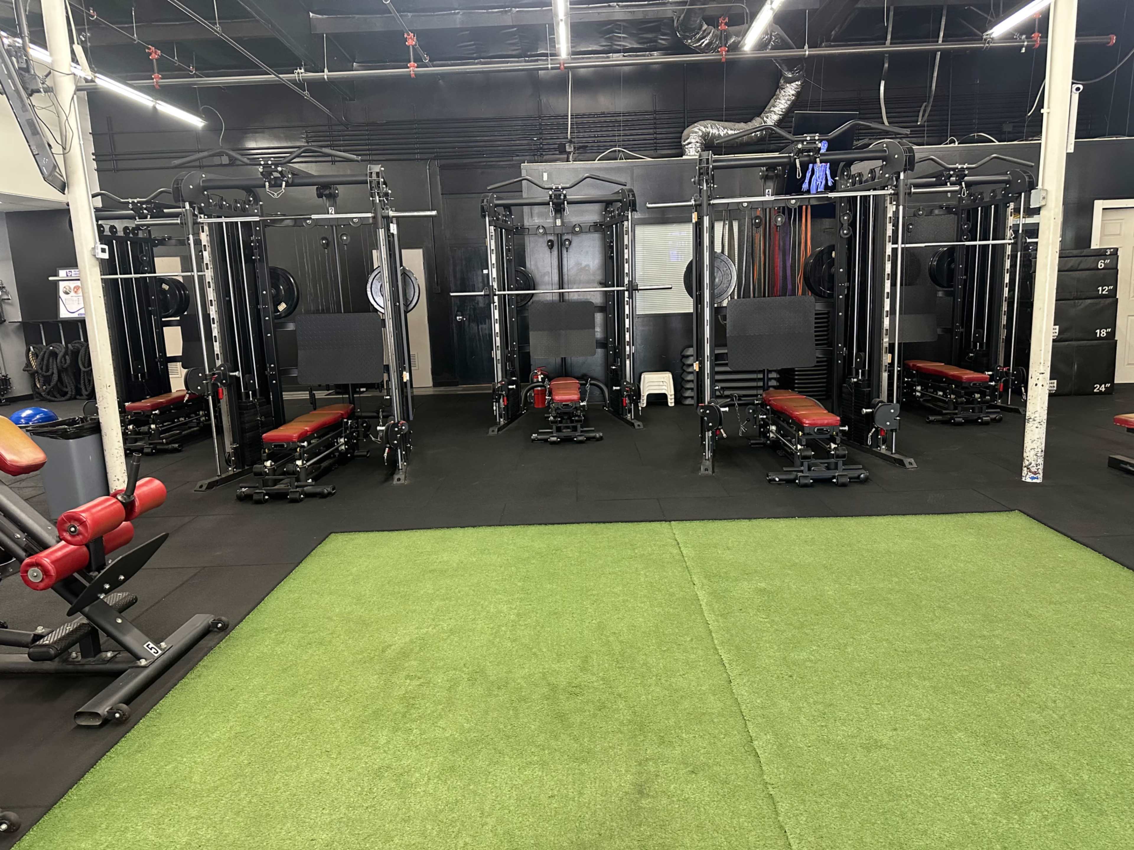 The image shows a gym interior equipped with multiple weight training machines arranged on a rubber floor with sections of green artificial turf.