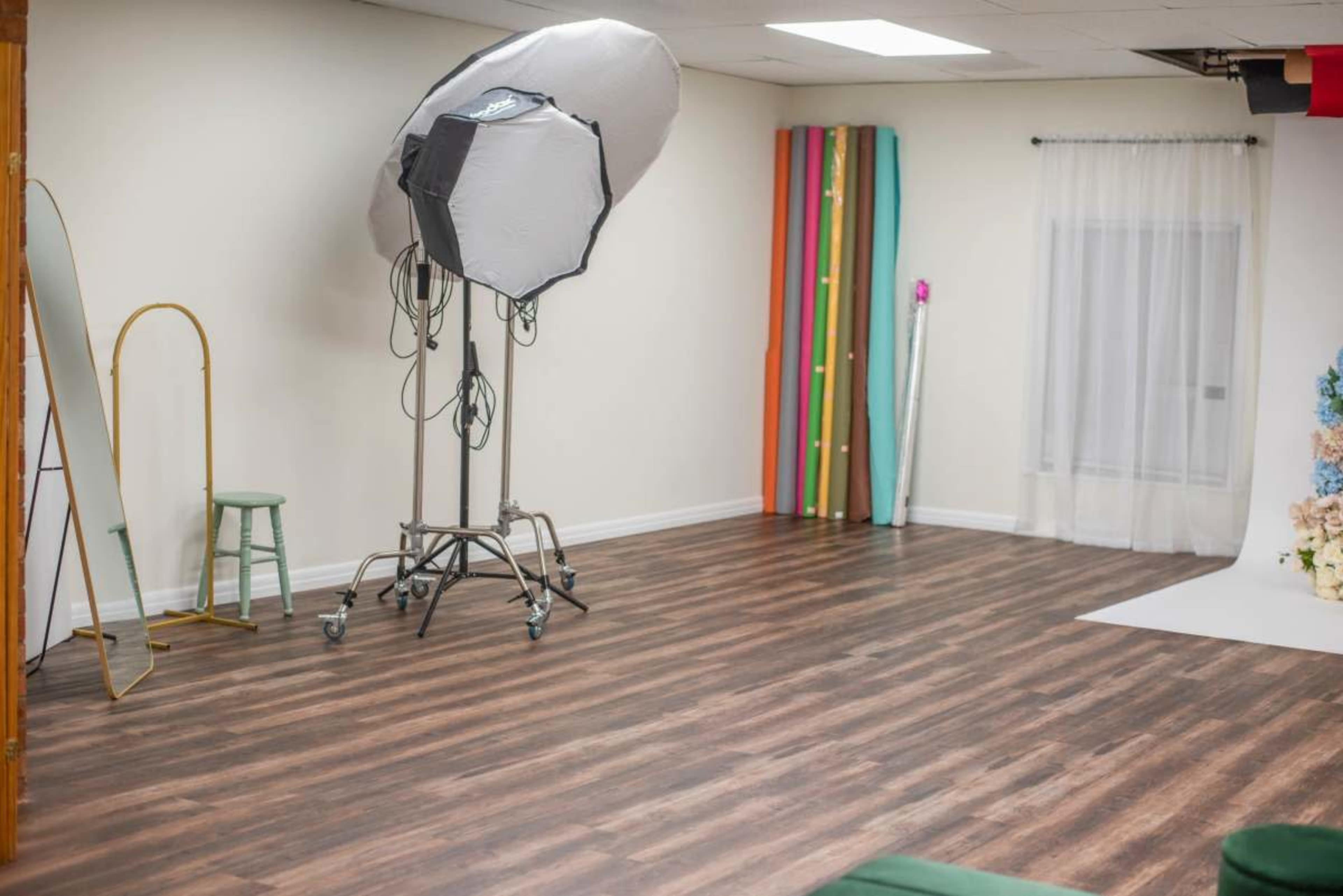 The image shows a well-lit photography studio with a large softbox light, a mirror, a small green stool, and colorful backdrops leaning against the wall.