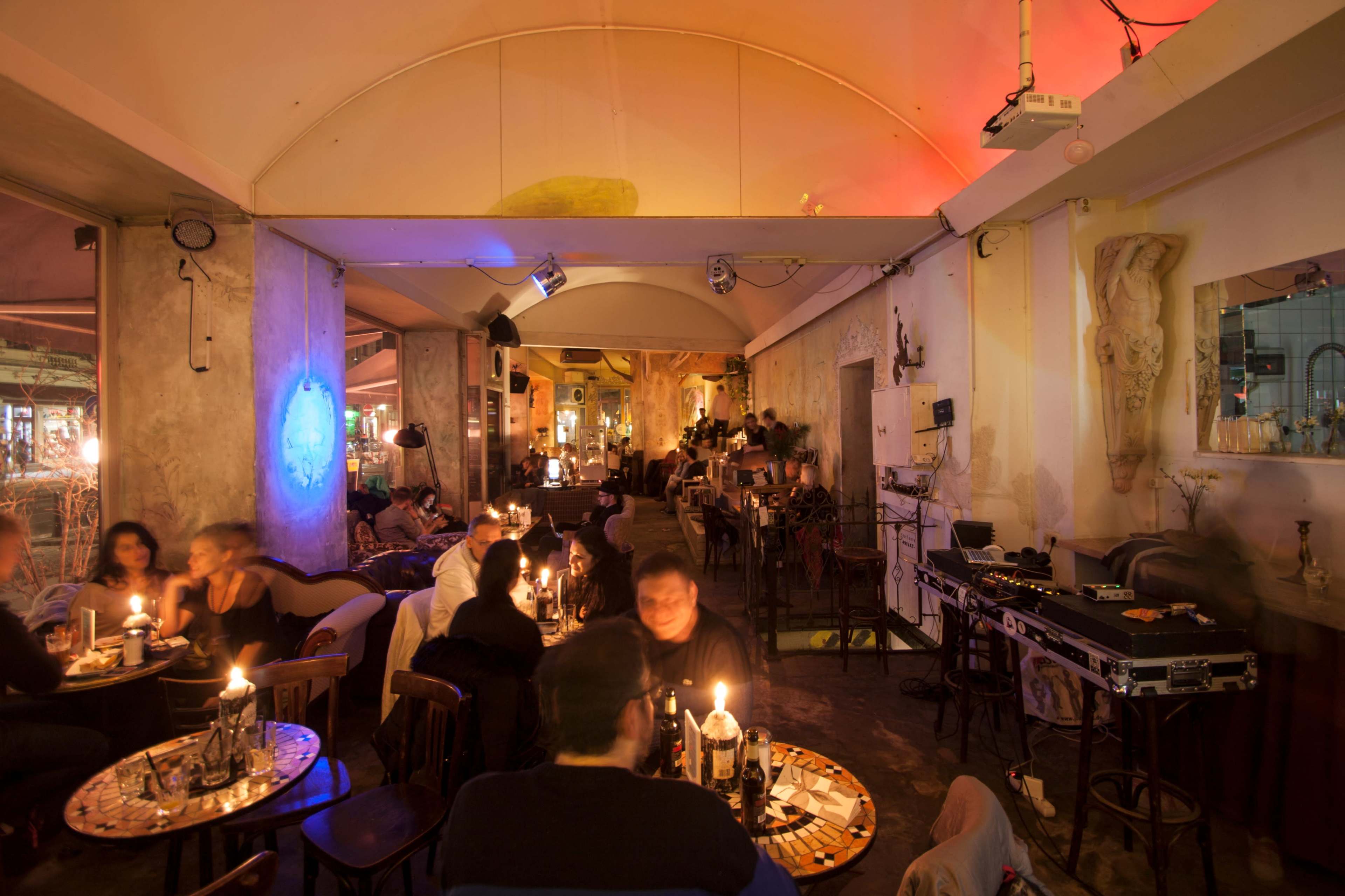 A dimly lit café features small tables with candlelight and a vibrant atmosphere filled with patrons enjoying their drinks and conversation.