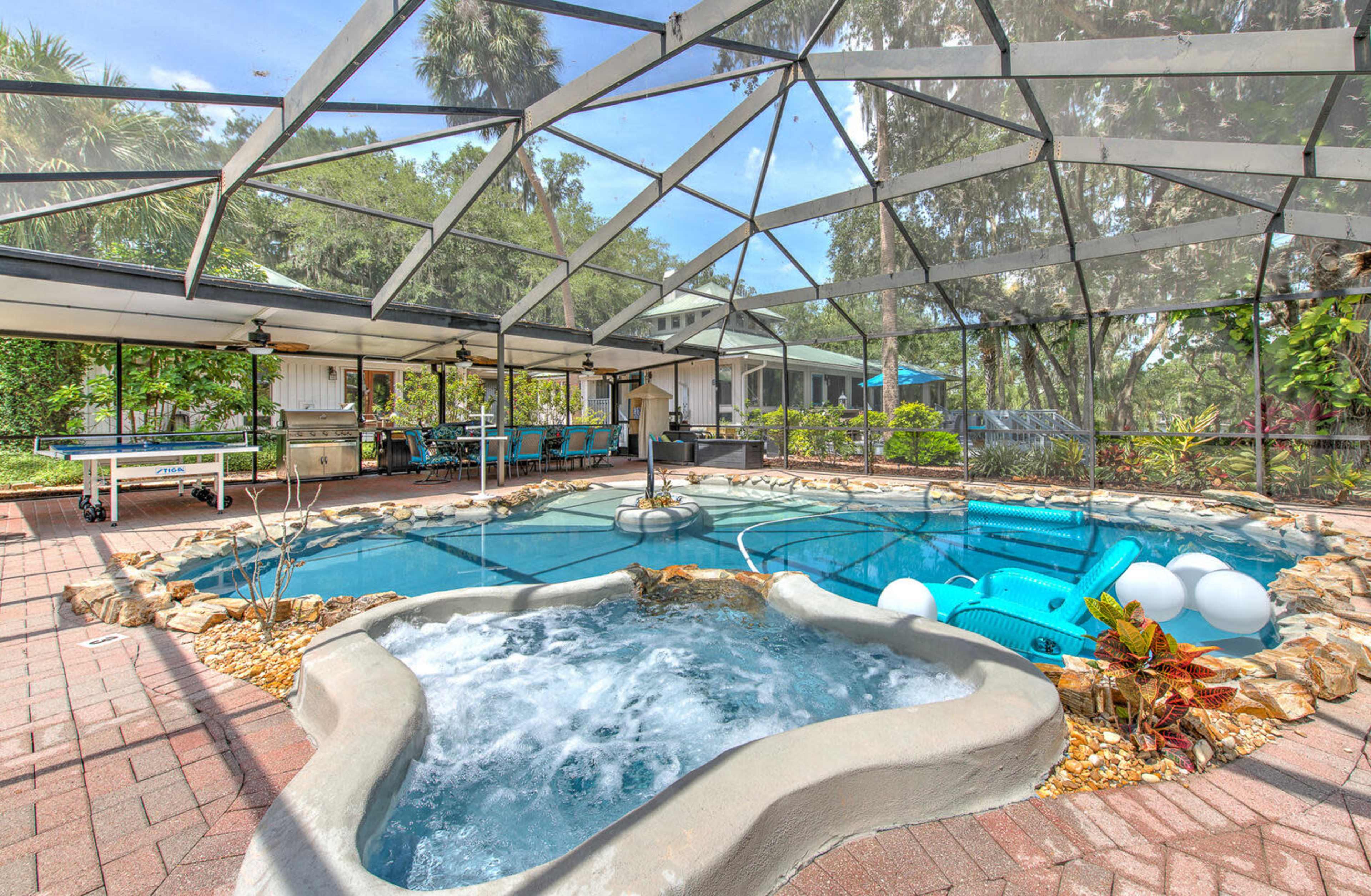 A screened-in pool area featuring a hot tub, swimming pool, and lounge seating surrounded by greenery.