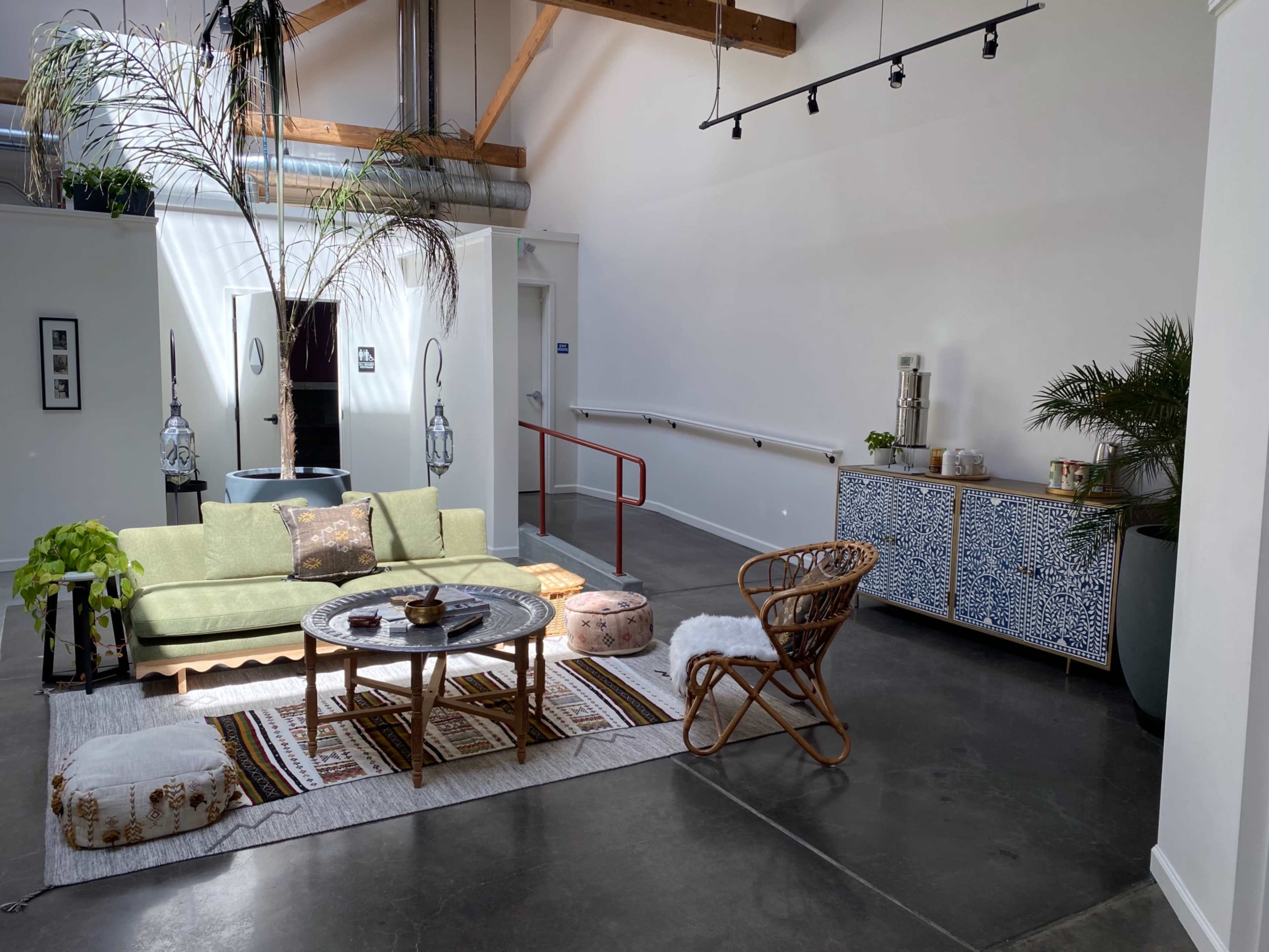 A living space features a green couch, round coffee table, patterned rug, and a woven chair, complemented by a decorative cabinet and potted plants.