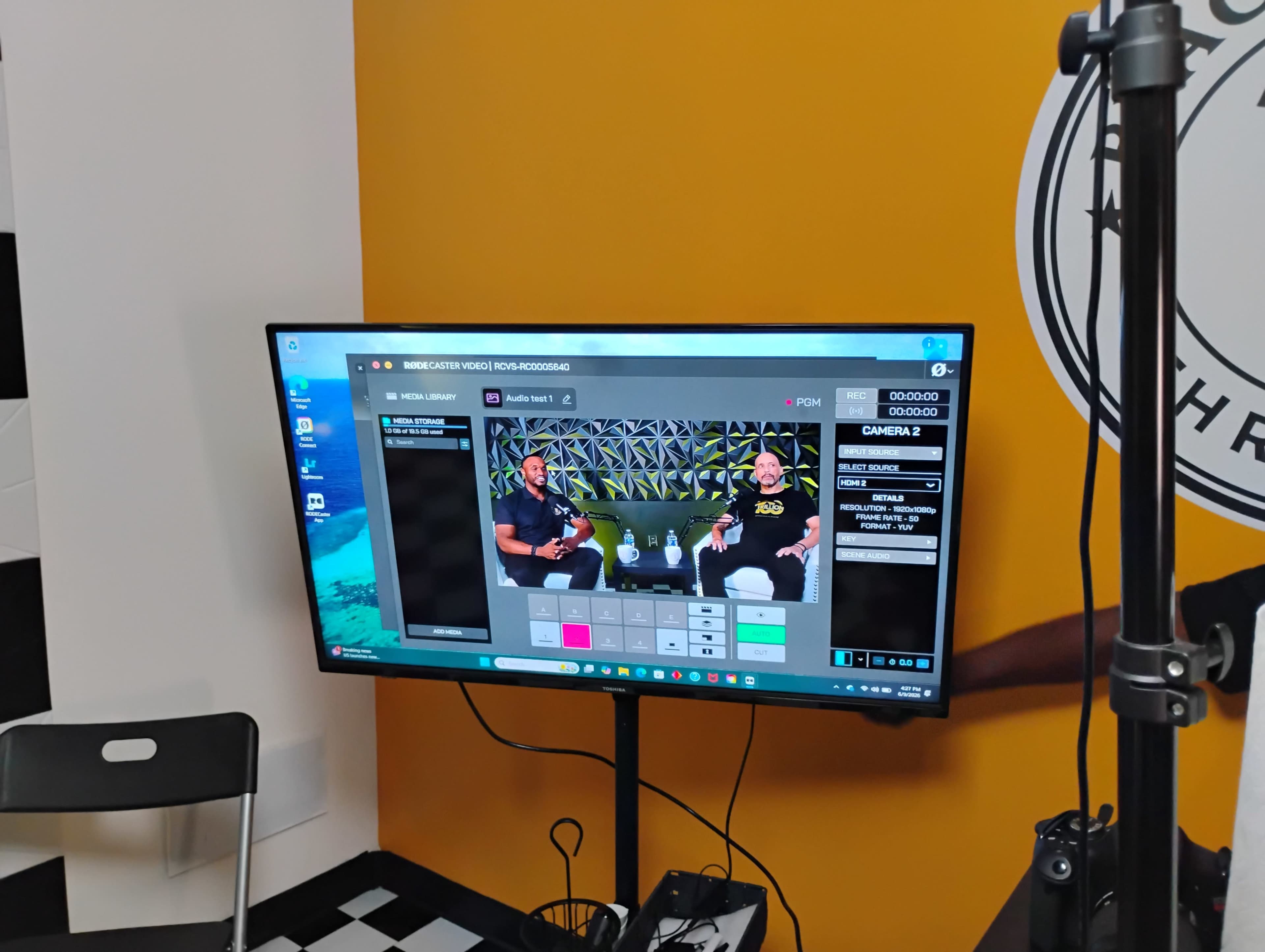 A television screen displays a video editing interface with two individuals seated and being recorded, set against a colorful background.