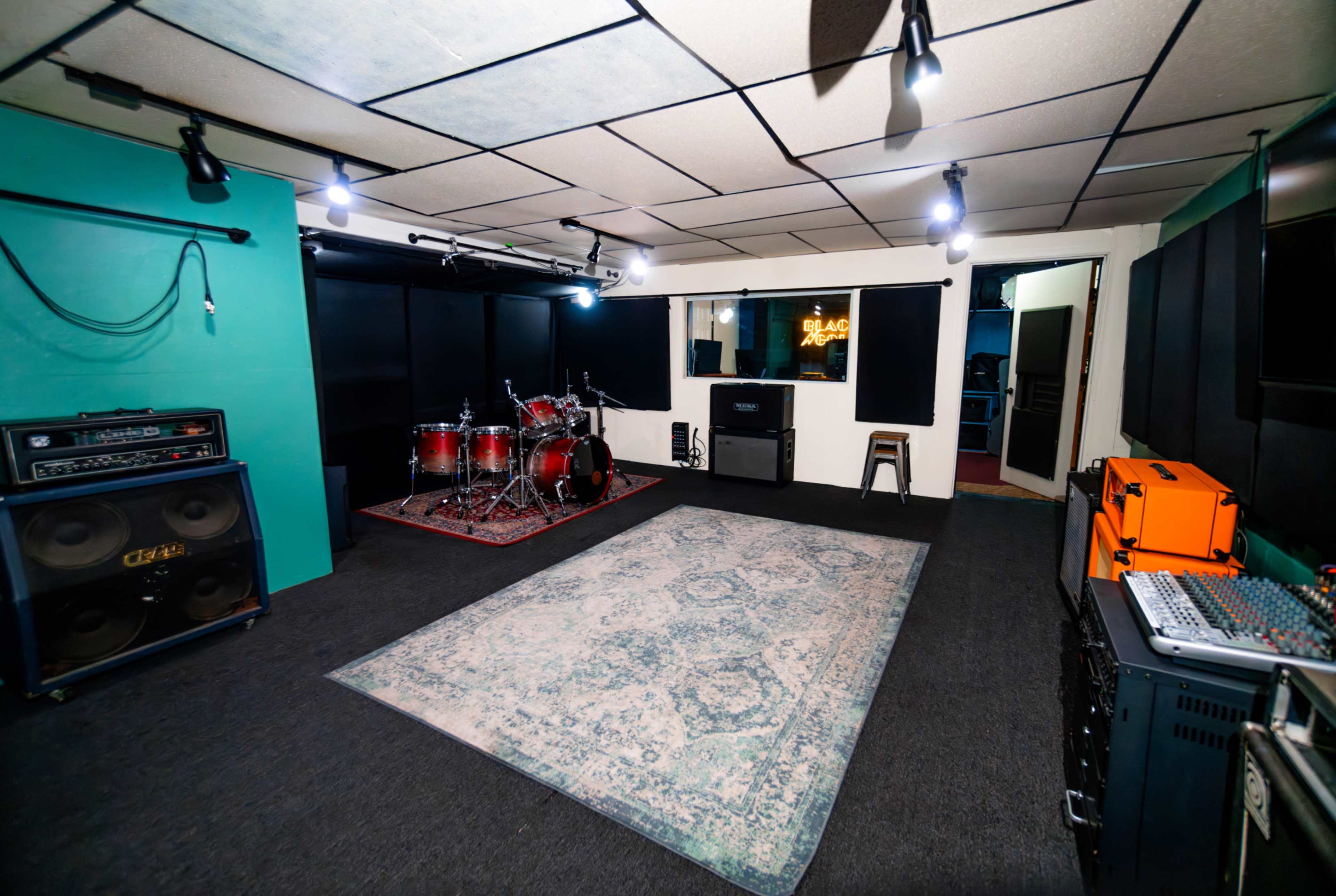 A music rehearsal studio with a drum set, amplifiers, and sound equipment arranged on a carpeted floor.