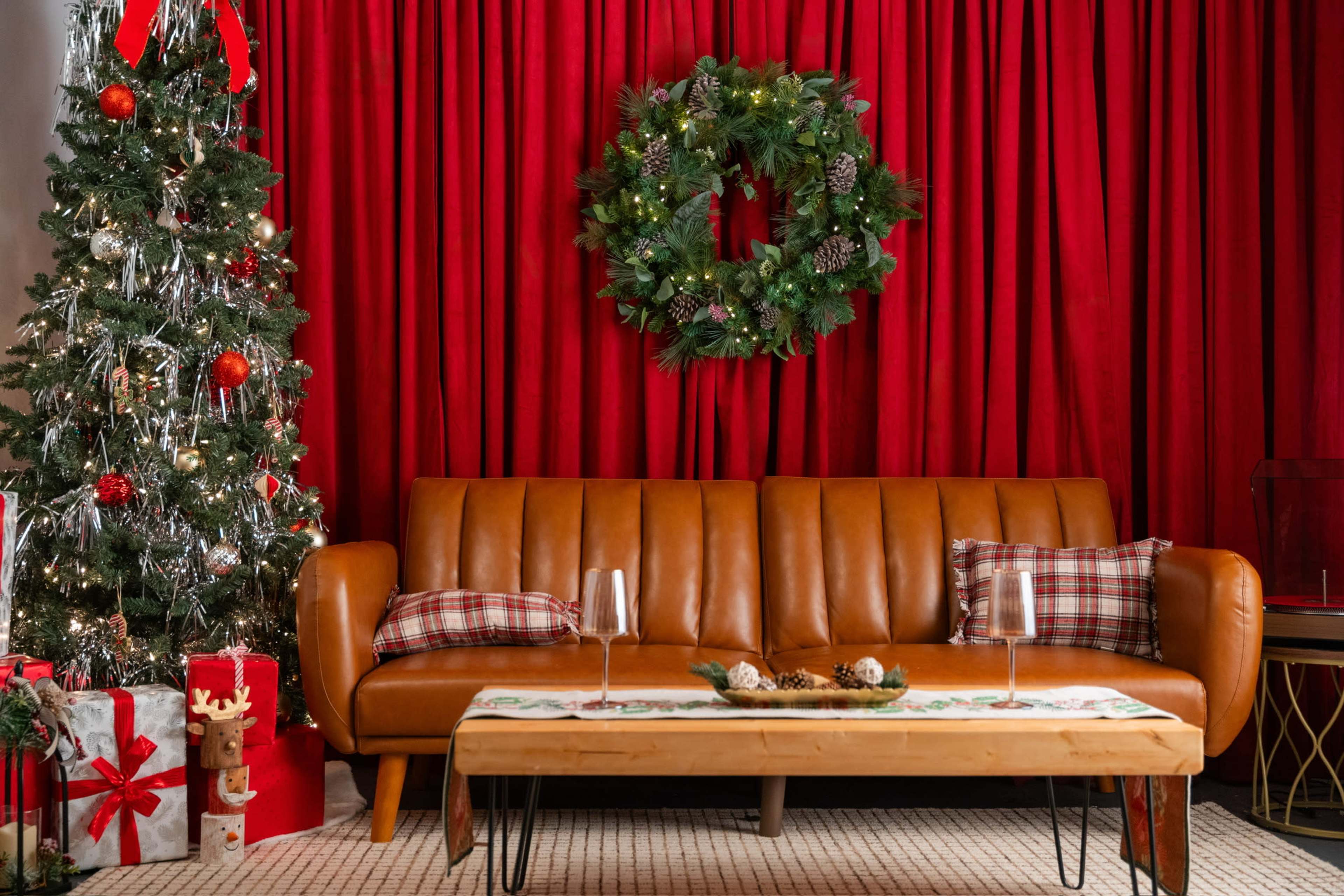 A brown leather sofa is positioned in front of a red curtain, decorated with a Christmas wreath, alongside a Christmas tree and wrapped gifts.