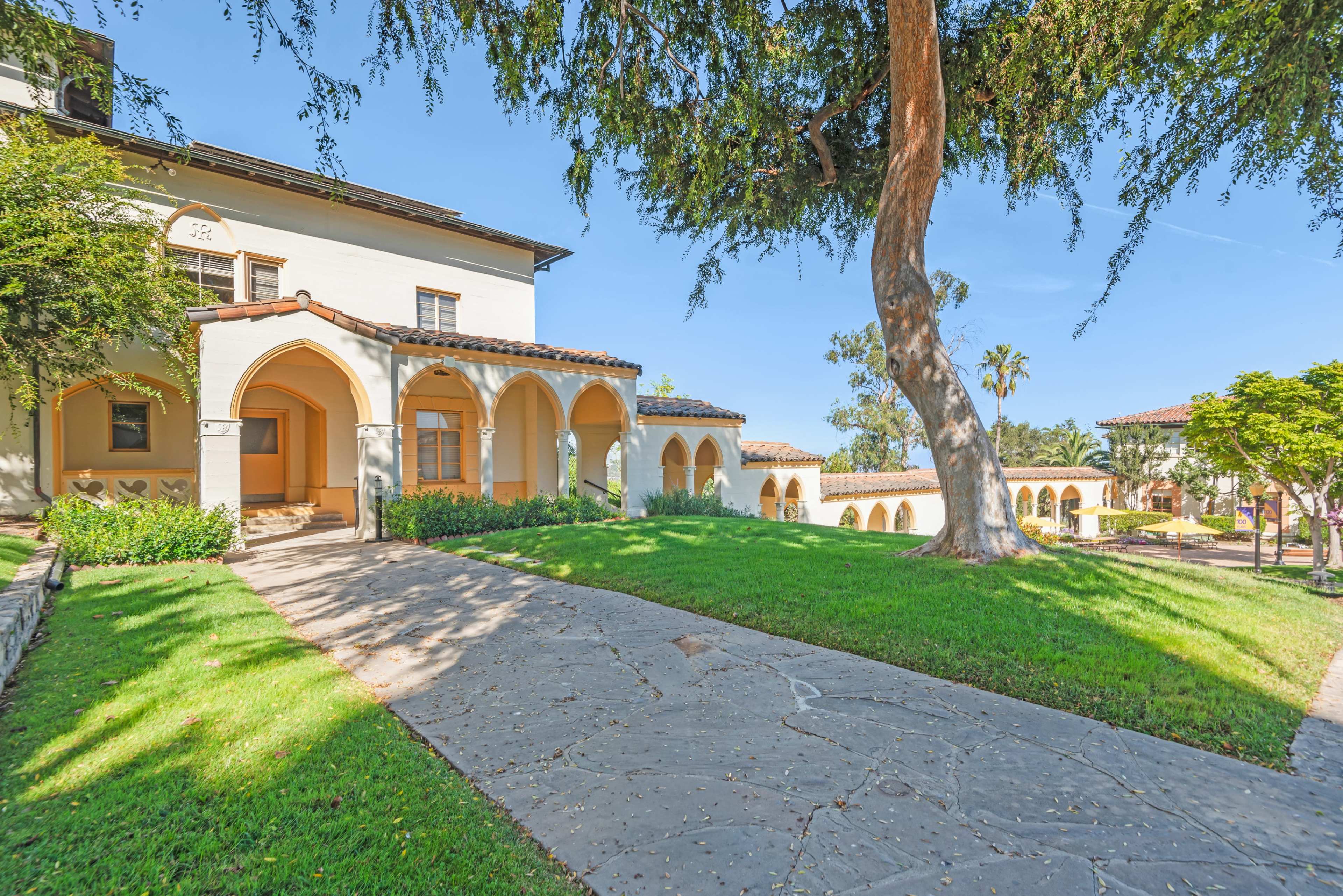 The image shows a Spanish-style building with arched doorways and a landscaped lawn featuring trees and shrubs.