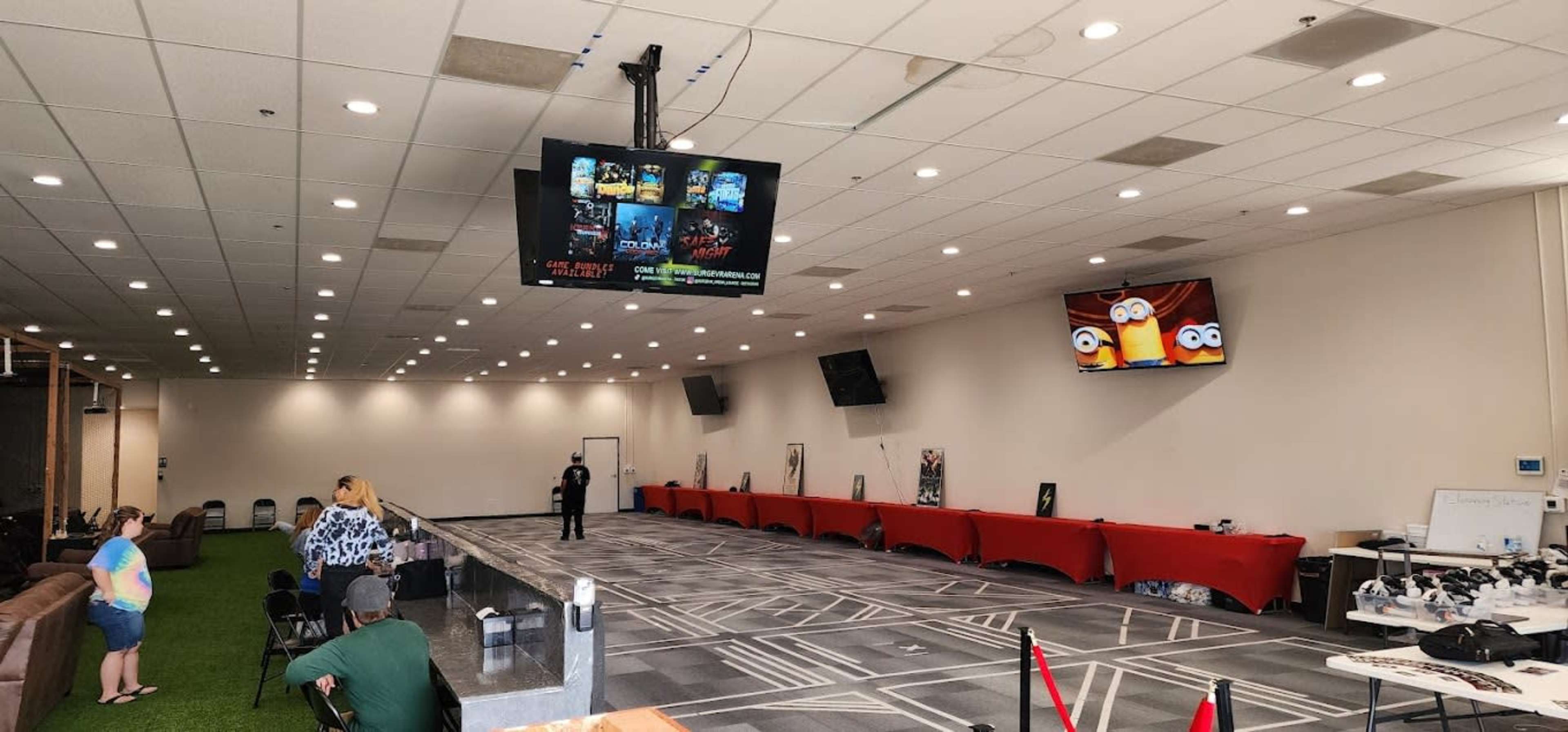 The image shows a spacious indoor event area with a carpeted floor, seating along the walls, tables covered in red cloths, and two screens displaying video content.