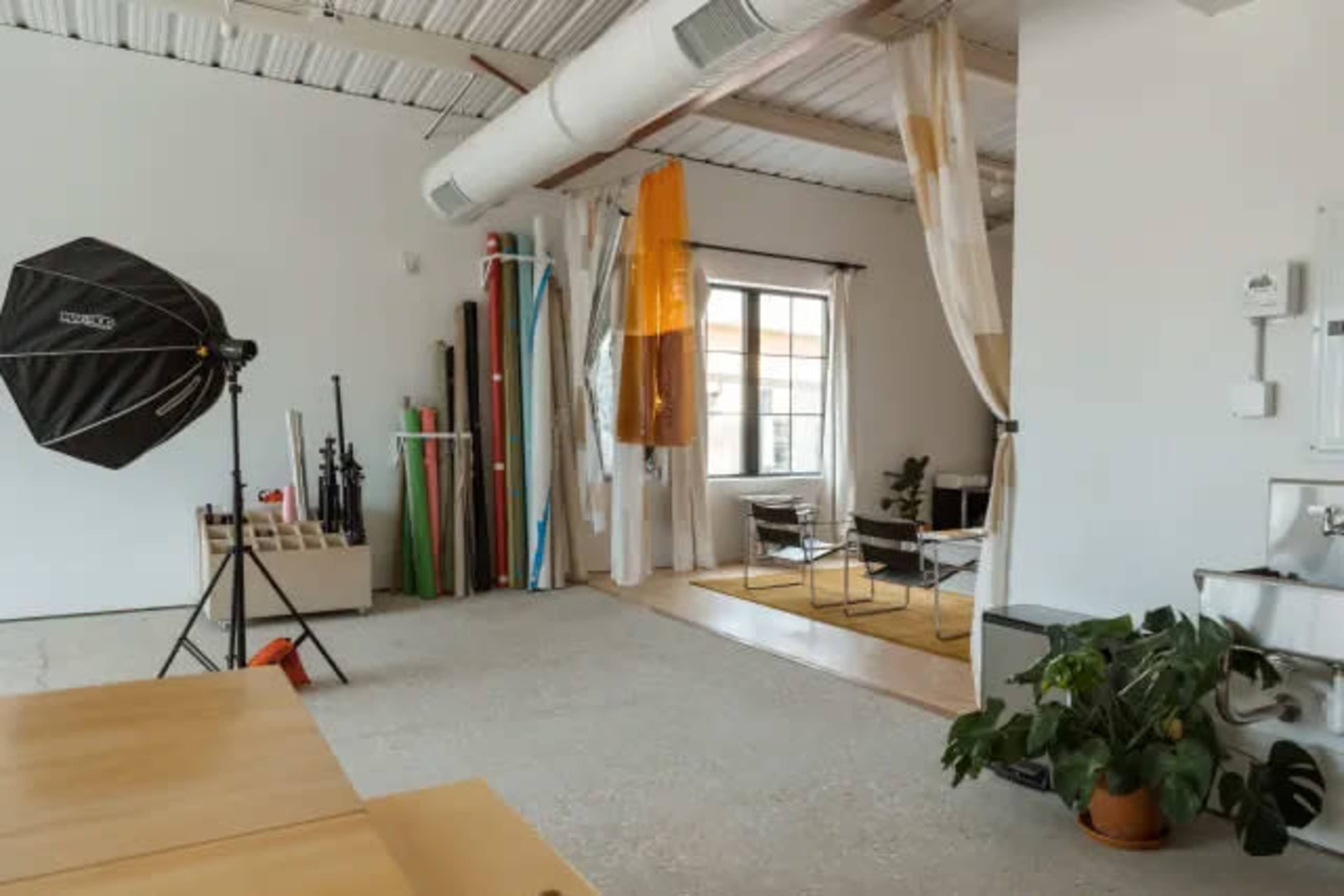 The image shows a spacious, well-lit studio with a photography setup, several background props, and a seating area.