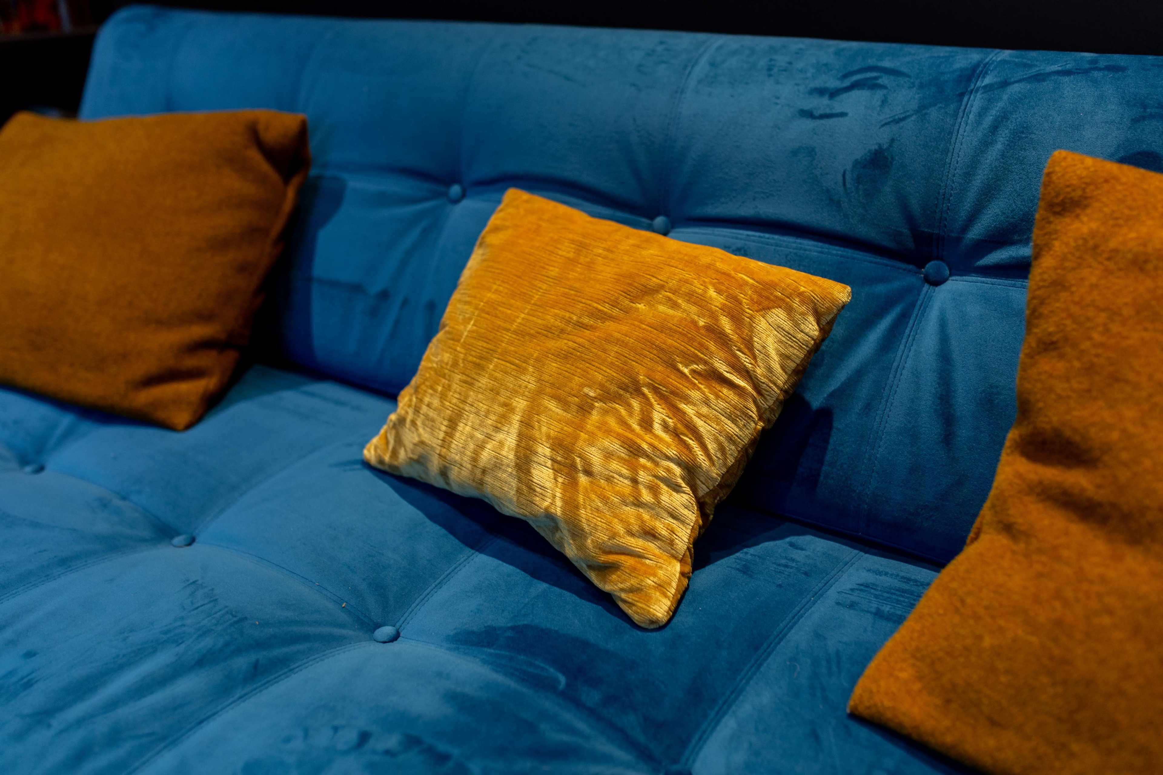 A blue velvet couch is adorned with two mustard-colored pillows, one of which is textured and prominently placed.
