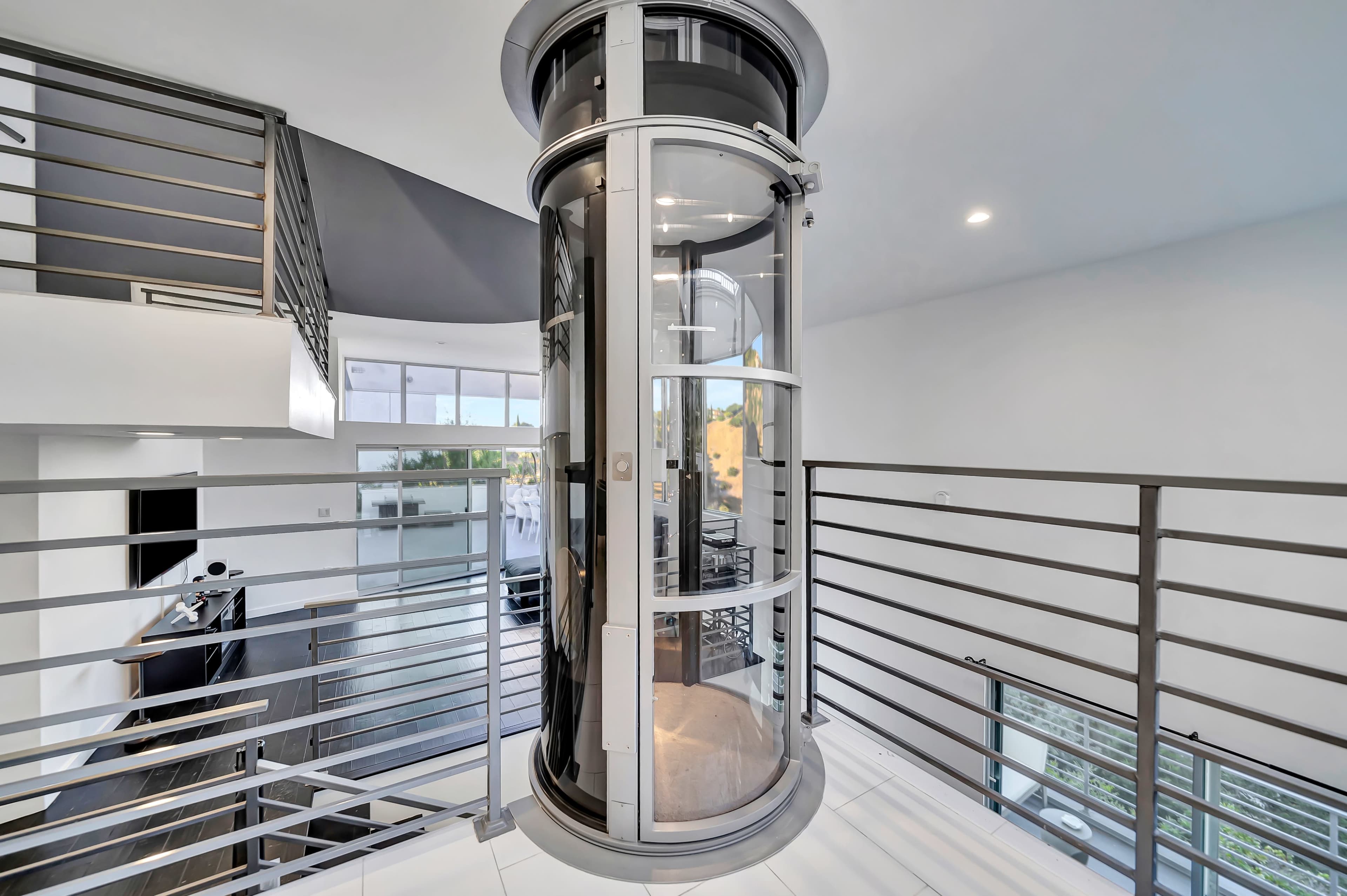 A modern glass elevator stands in a spacious, contemporary home surrounded by metal railings and an open interior.