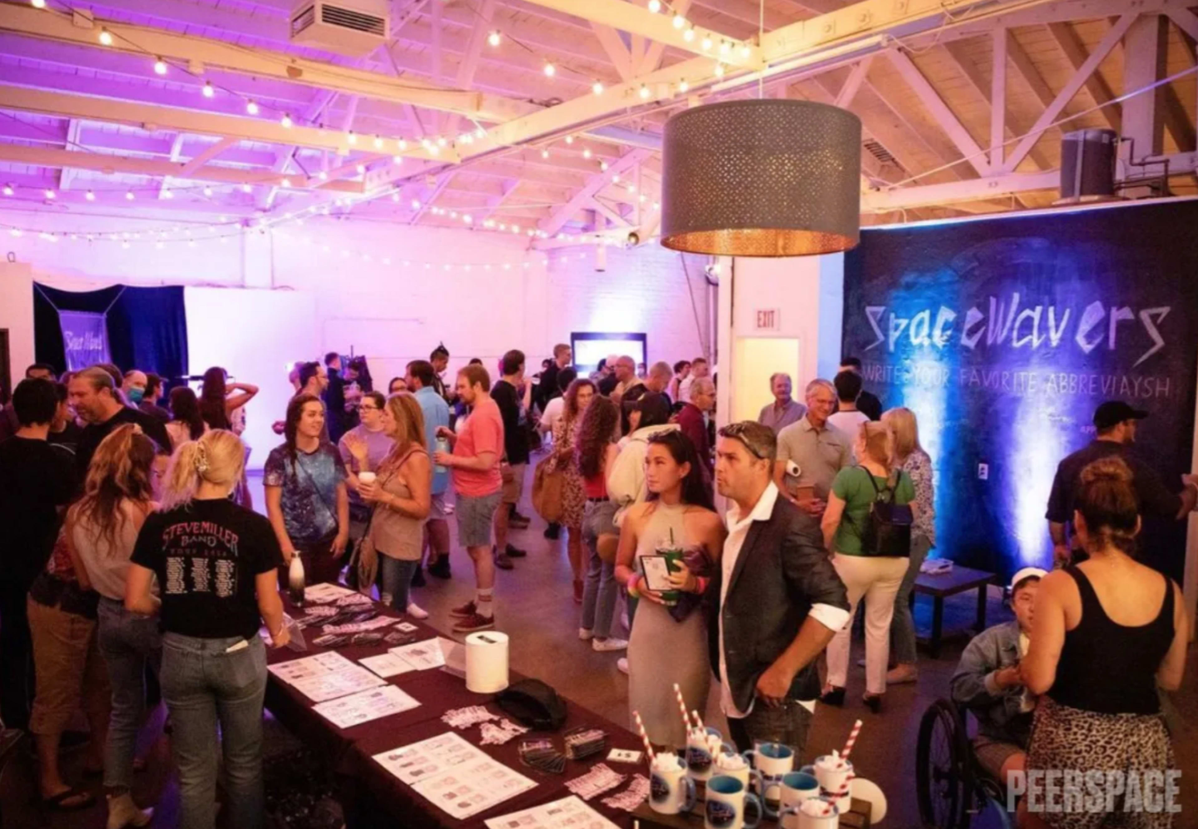 A large crowd gathers in a well-lit venue decorated with string lights, engaging in conversation and networking around tables filled with various promotional materials.