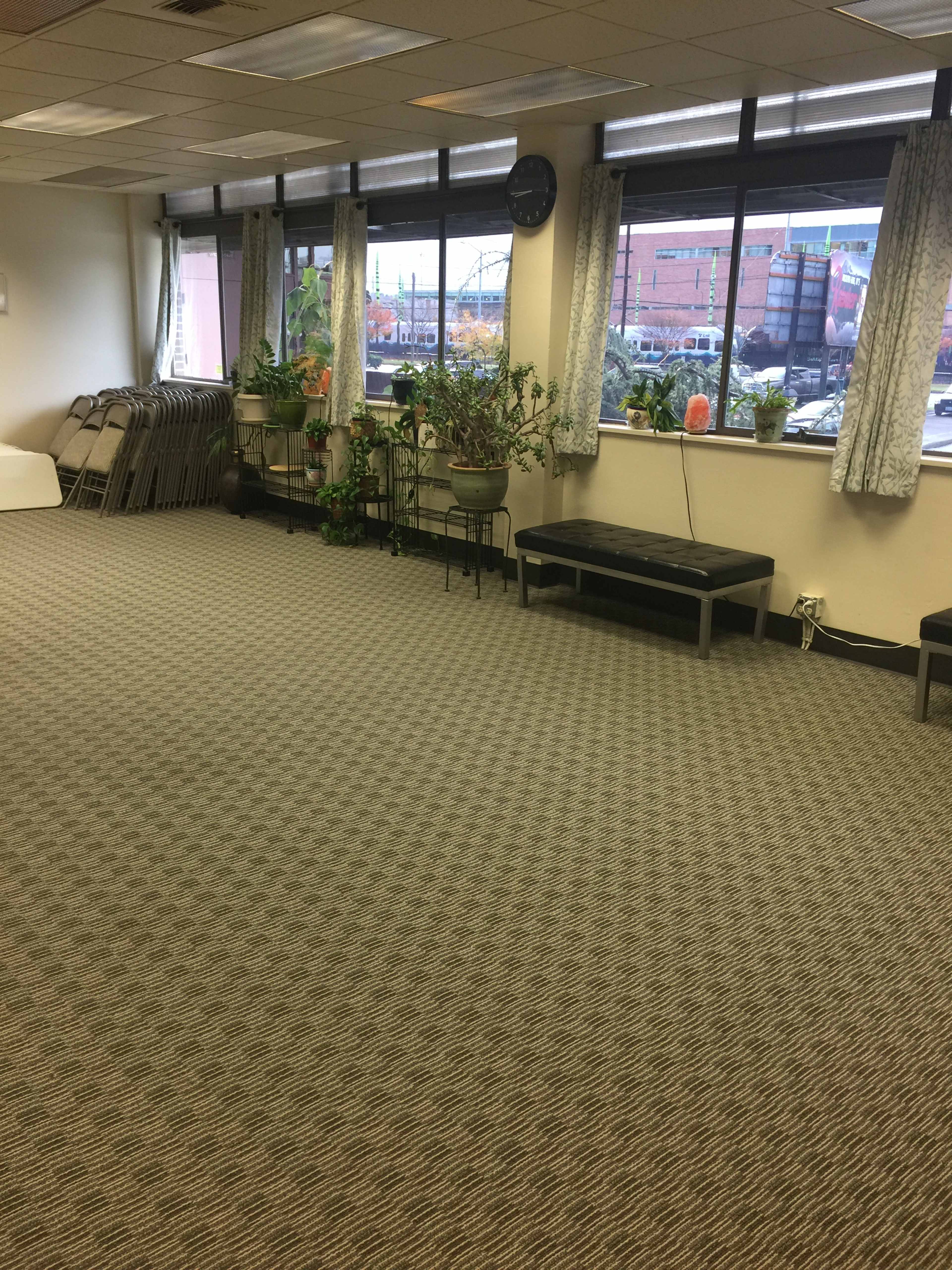 An empty office space with a carpeted floor, several chairs stacked against the wall, and plants near the windows.