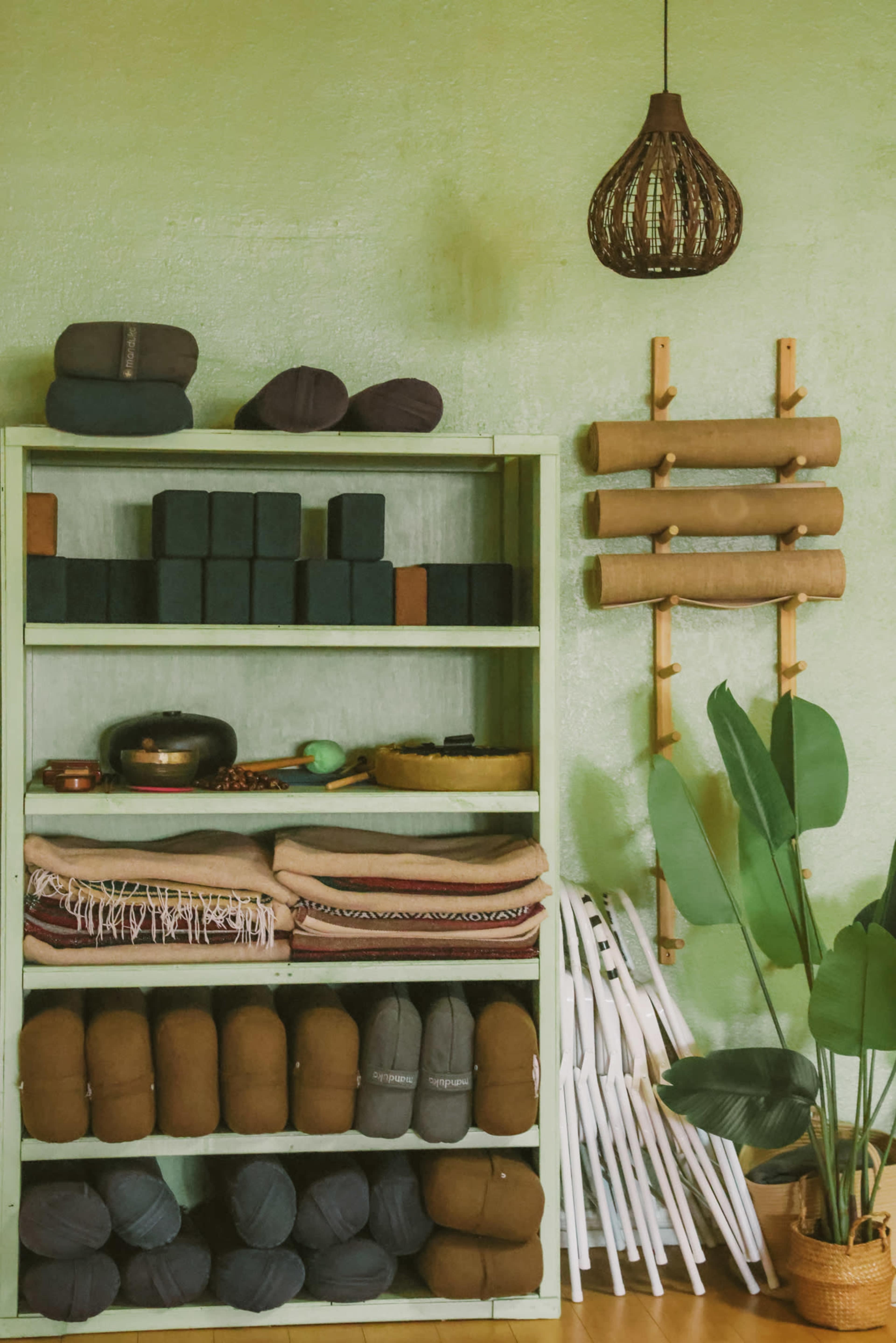 A green wall features a wooden shelving unit filled with yoga props, including cushions, blocks, and blankets, alongside a tall plant and wall-mounted storage.