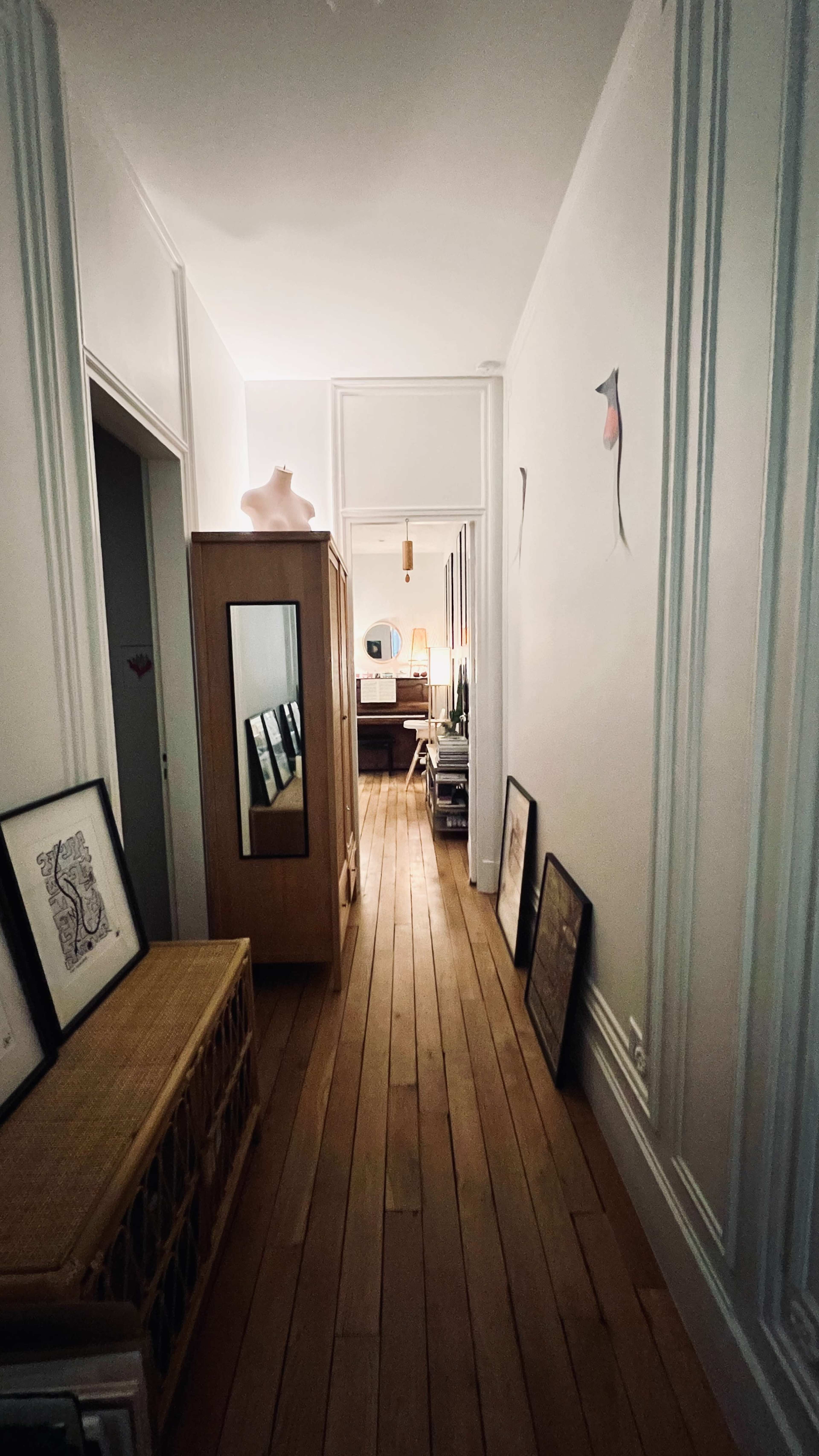 A narrow hallway features wooden flooring, framed artwork leaning against the walls, and a wooden cabinet on one side.