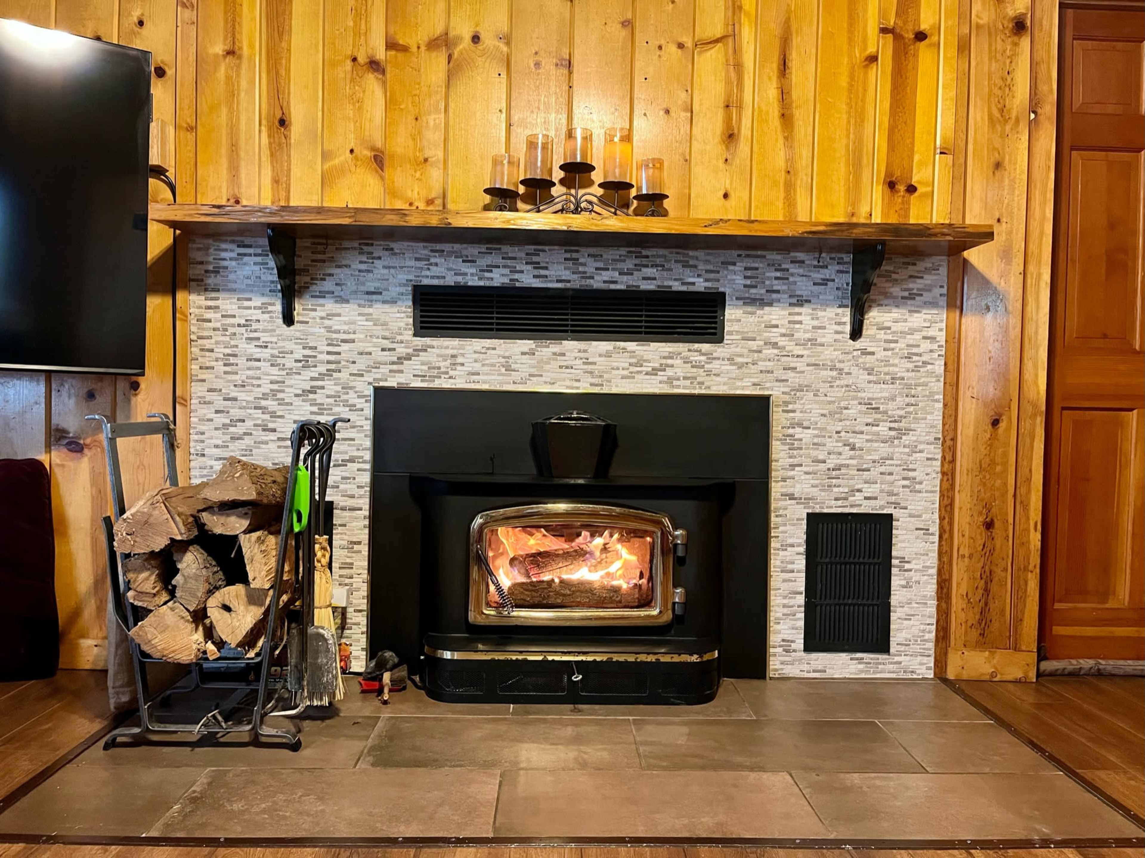 A wood-burning stove set in a stone facade, with a stack of firewood beside it and wooden paneling on the walls.