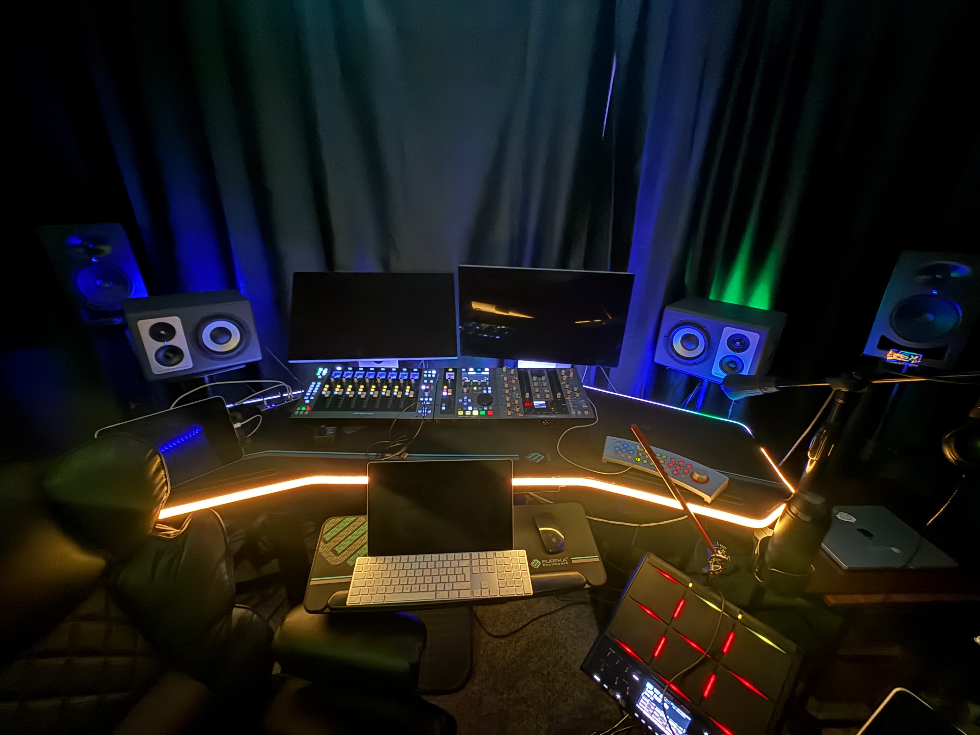 The image shows a music production studio with a computer setup, multiple monitors, audio interfaces, and various equipment, all surrounded by dark curtains and illuminated with colored lights.