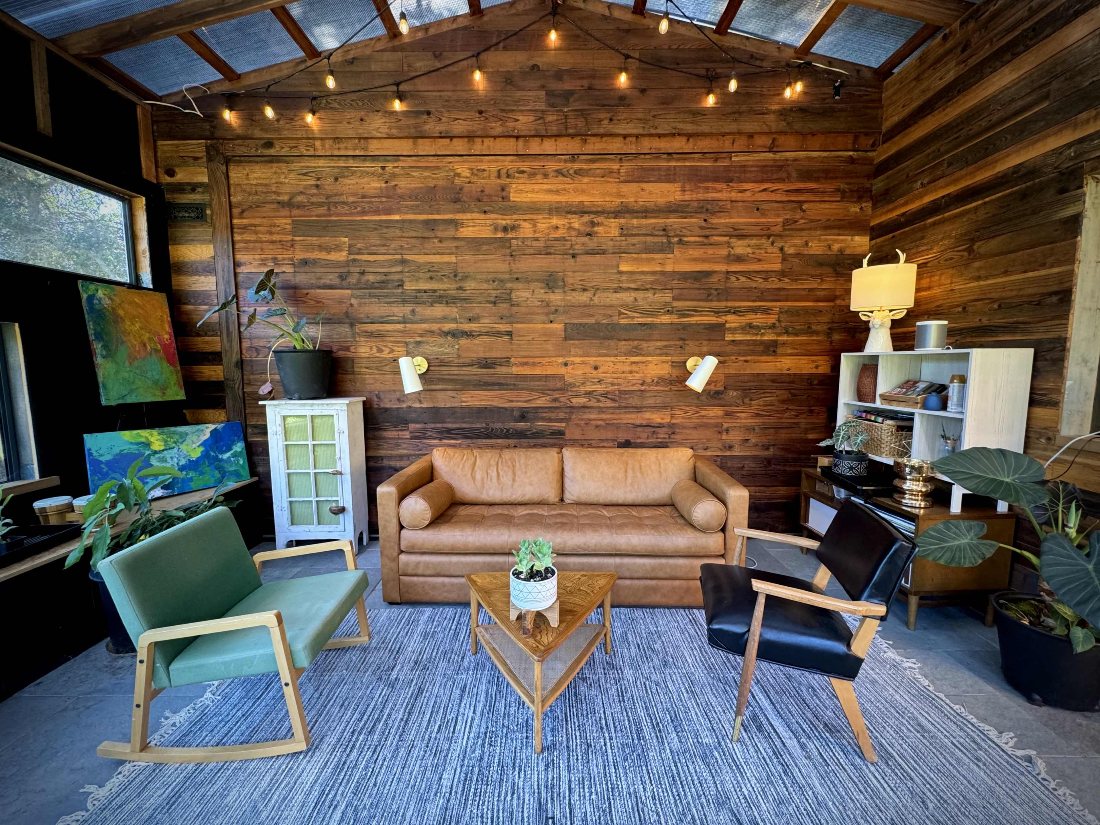 The image shows a cozy living space featuring a brown leather sofa, two armchairs, a wooden coffee table, and a textured rug against a wooden-paneled wall.