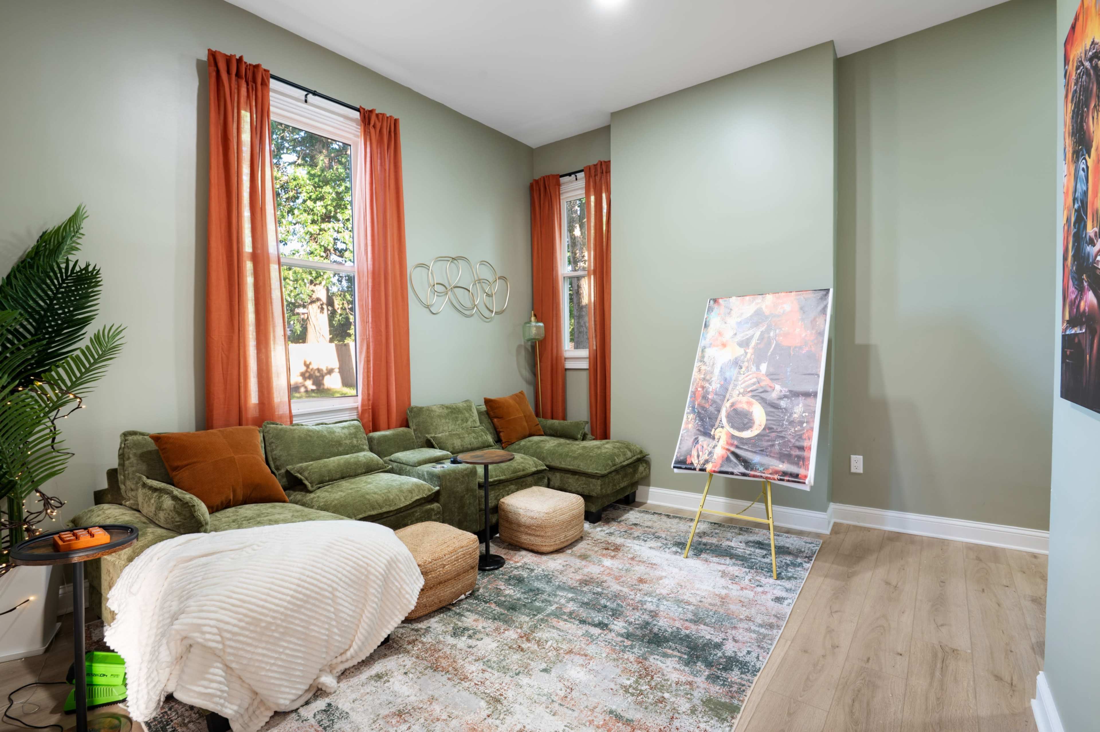 The image shows a cozy living room with a green sectional sofa, large windows with orange curtains, a decorative painting on an easel, and a textured area rug on a wooden floor.