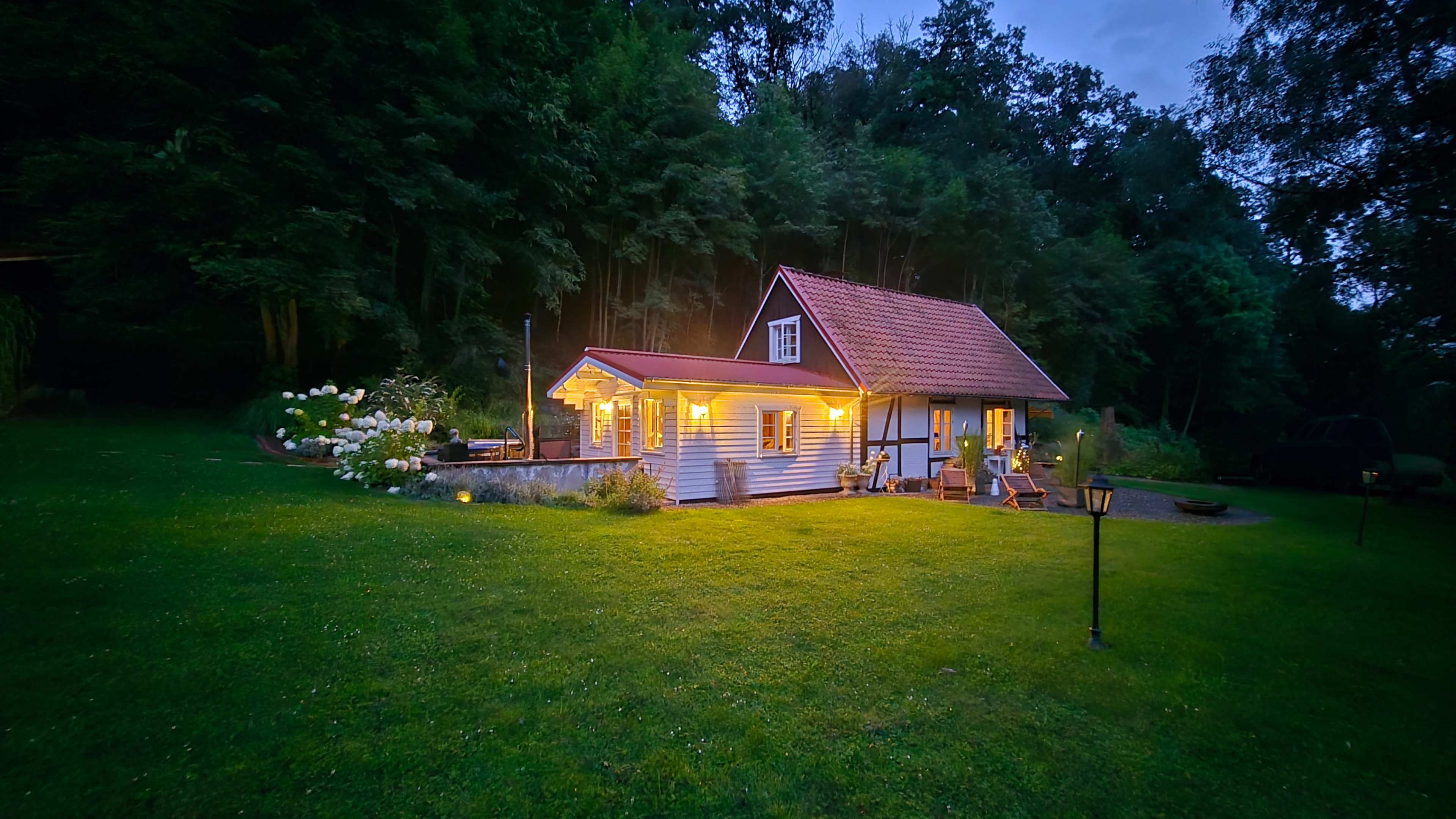 A wooden house with a red roof is lit up in the evening, surrounded by greenery and a well-maintained lawn.