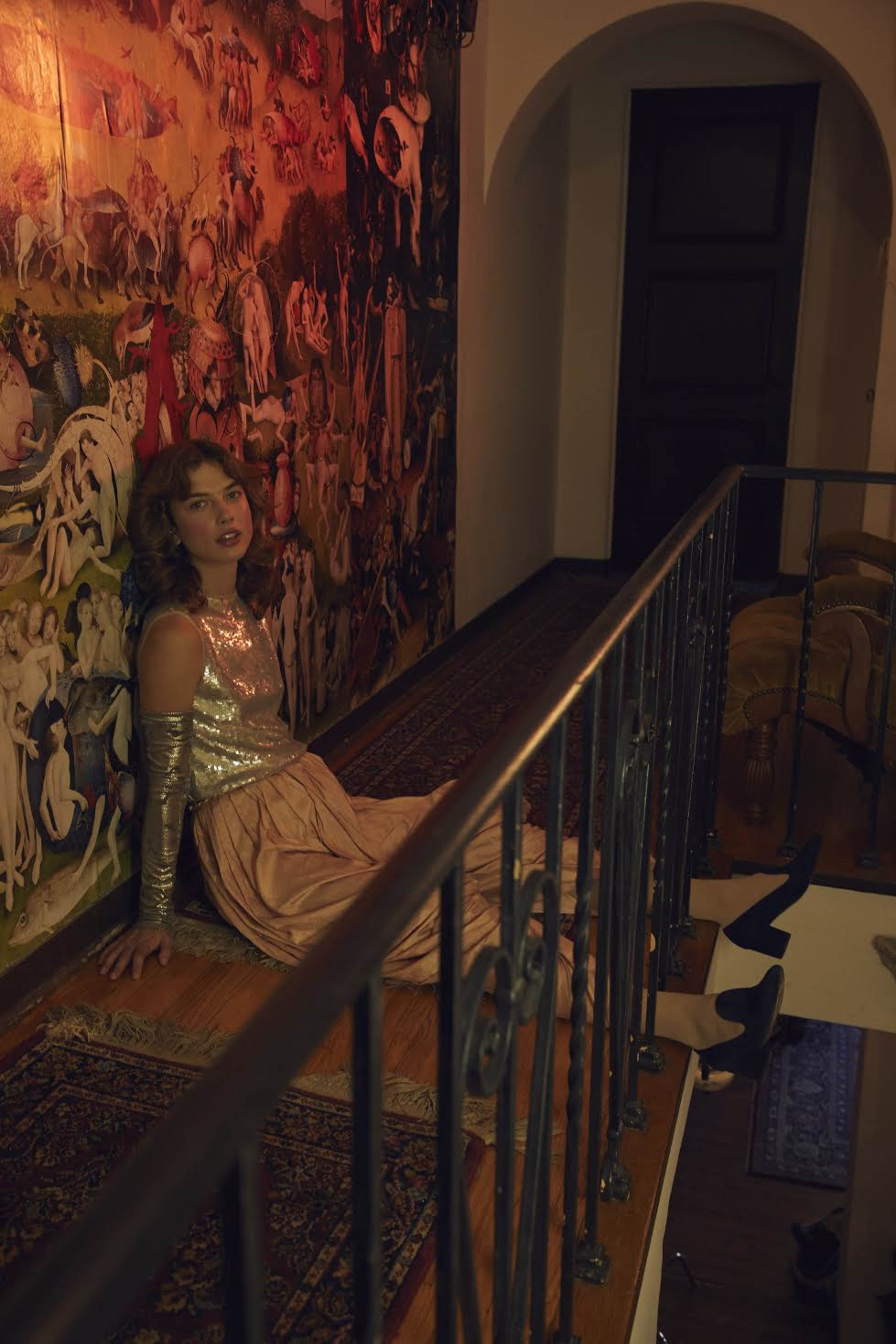 A woman in a sequined top and long skirt sits against a wall adorned with an elaborate mural, in a softly lit hallway.
