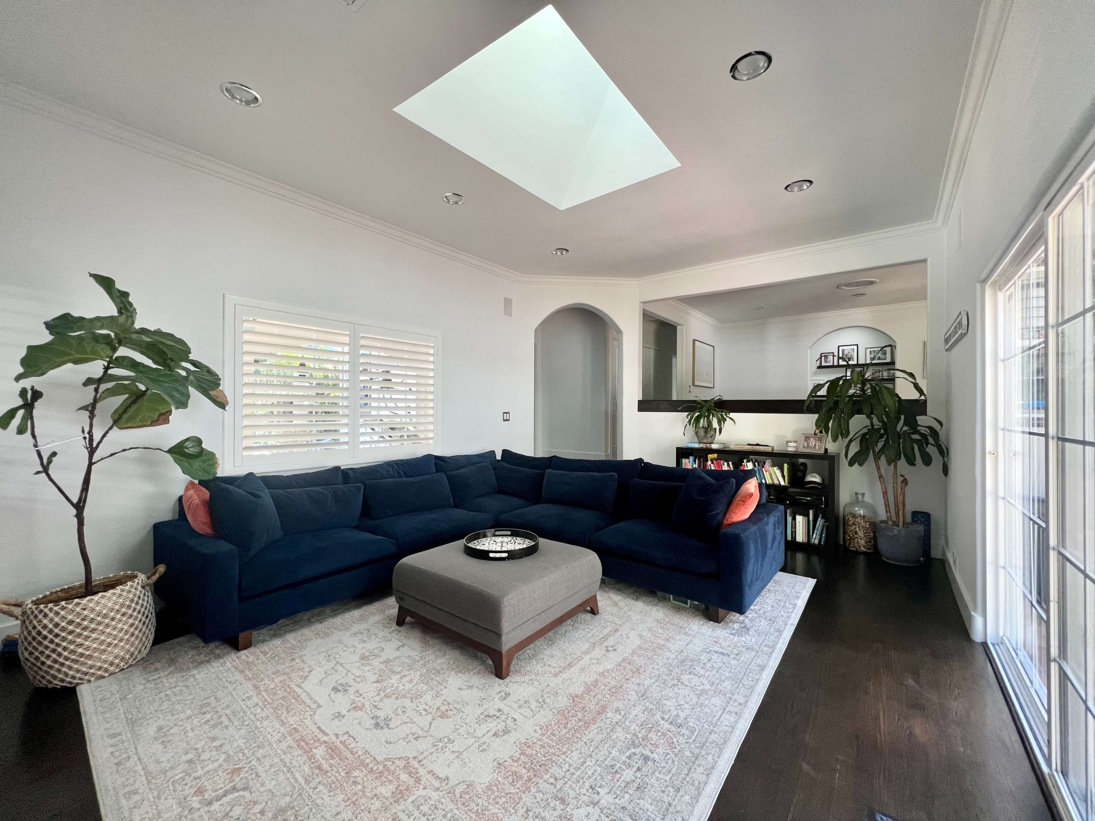 A spacious living room features a blue sectional sofa, a large area rug, and a skylight above, with plants and a small dining area visible in the background.