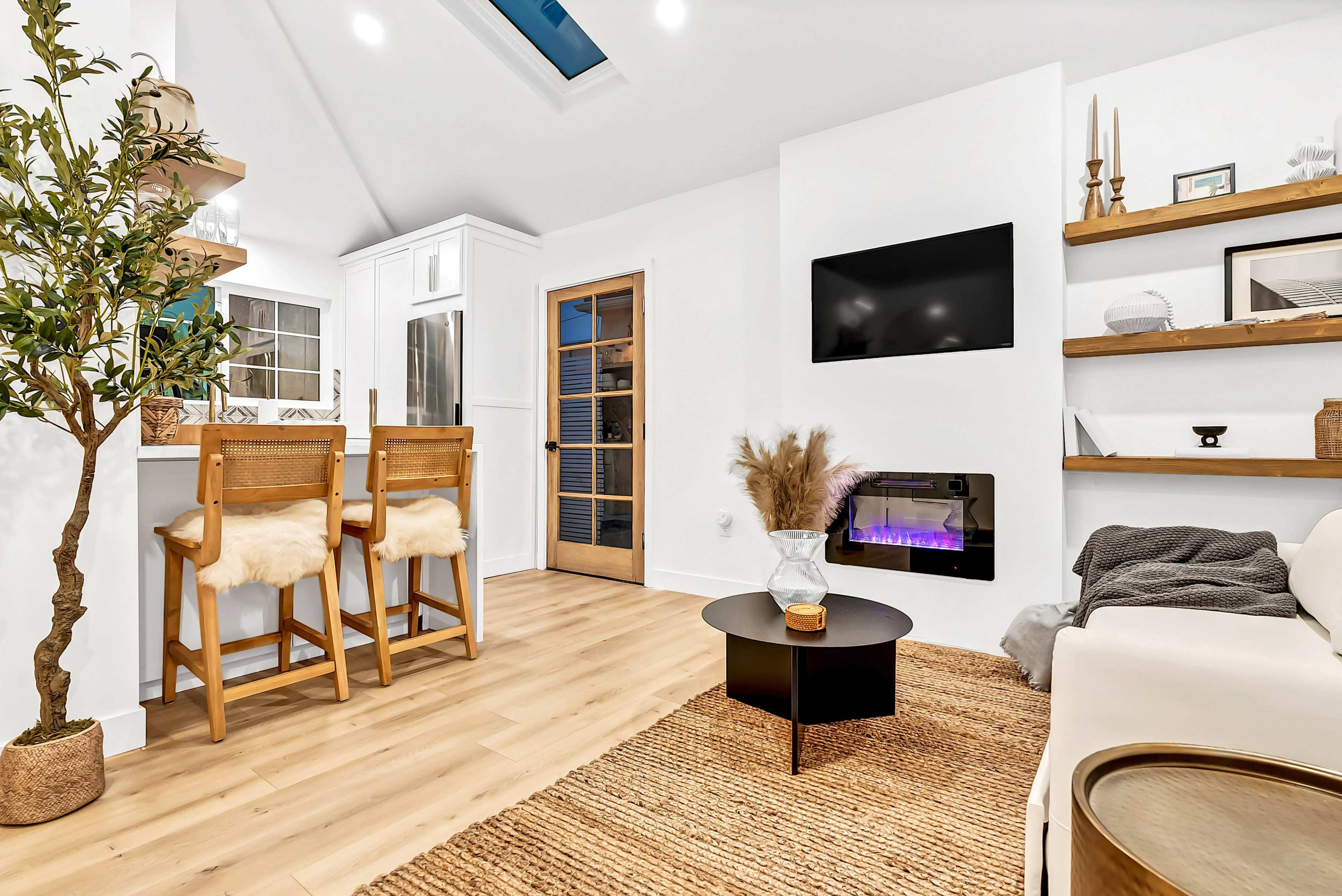 The image shows a modern living space with a kitchen bar featuring two stools, a wall-mounted TV, and a decorative shelf with plants and accessories.