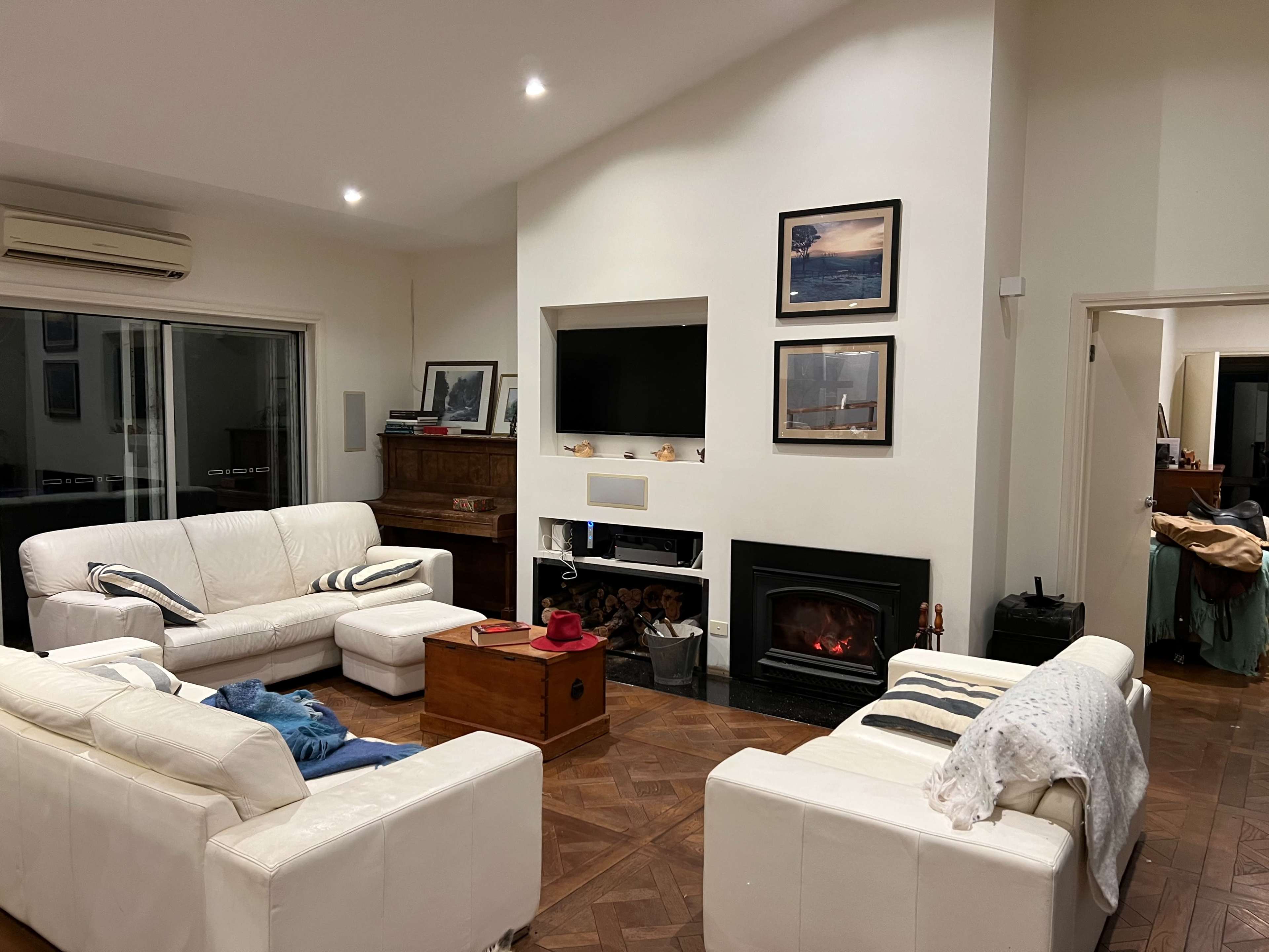 A spacious living room features white leather furniture arranged around a central coffee table and a fireplace, with framed artwork on the walls and a television mounted above the mantel.