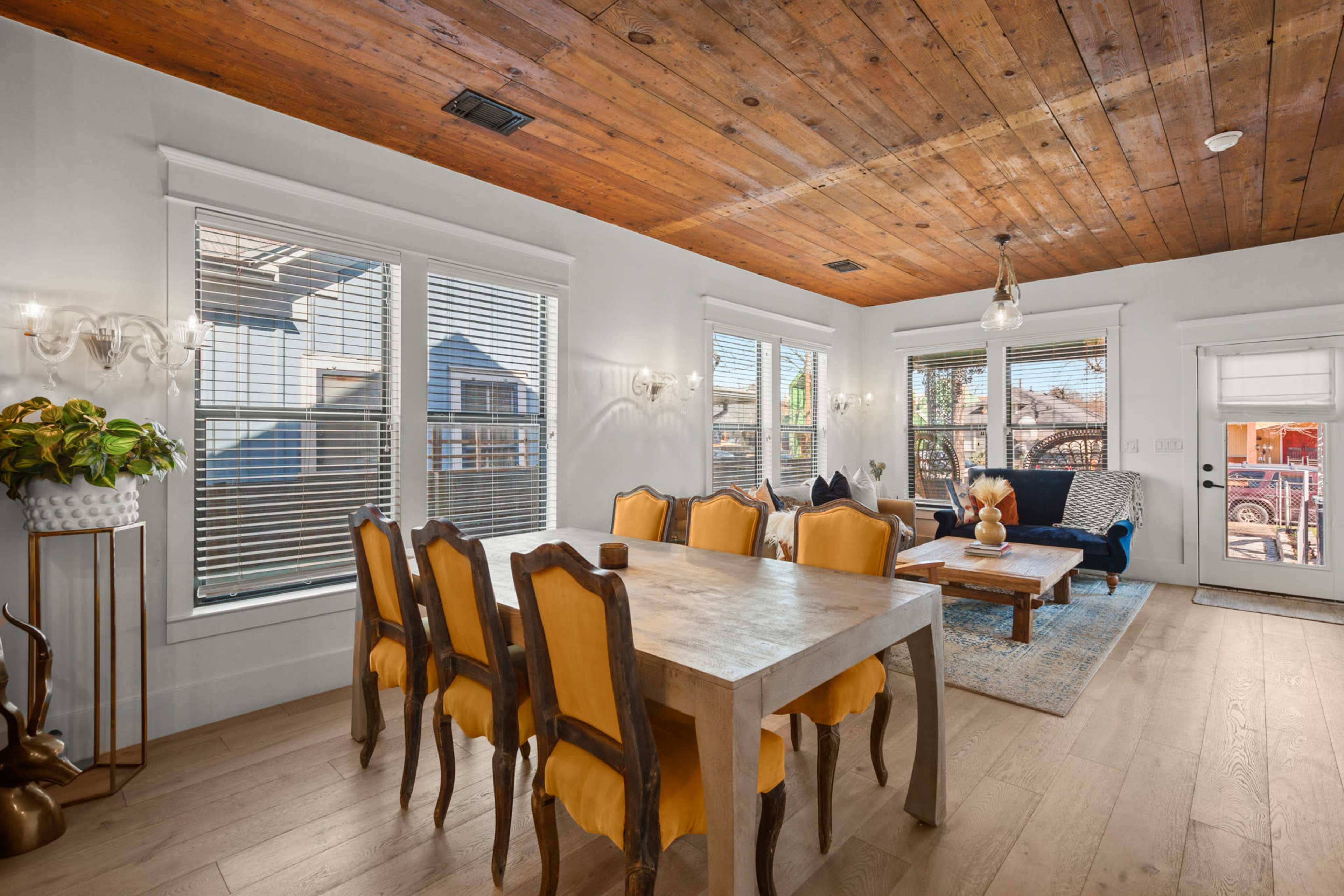 A dining area features a wooden table surrounded by yellow upholstered chairs, with large windows providing natural light and a view of the outdoors.