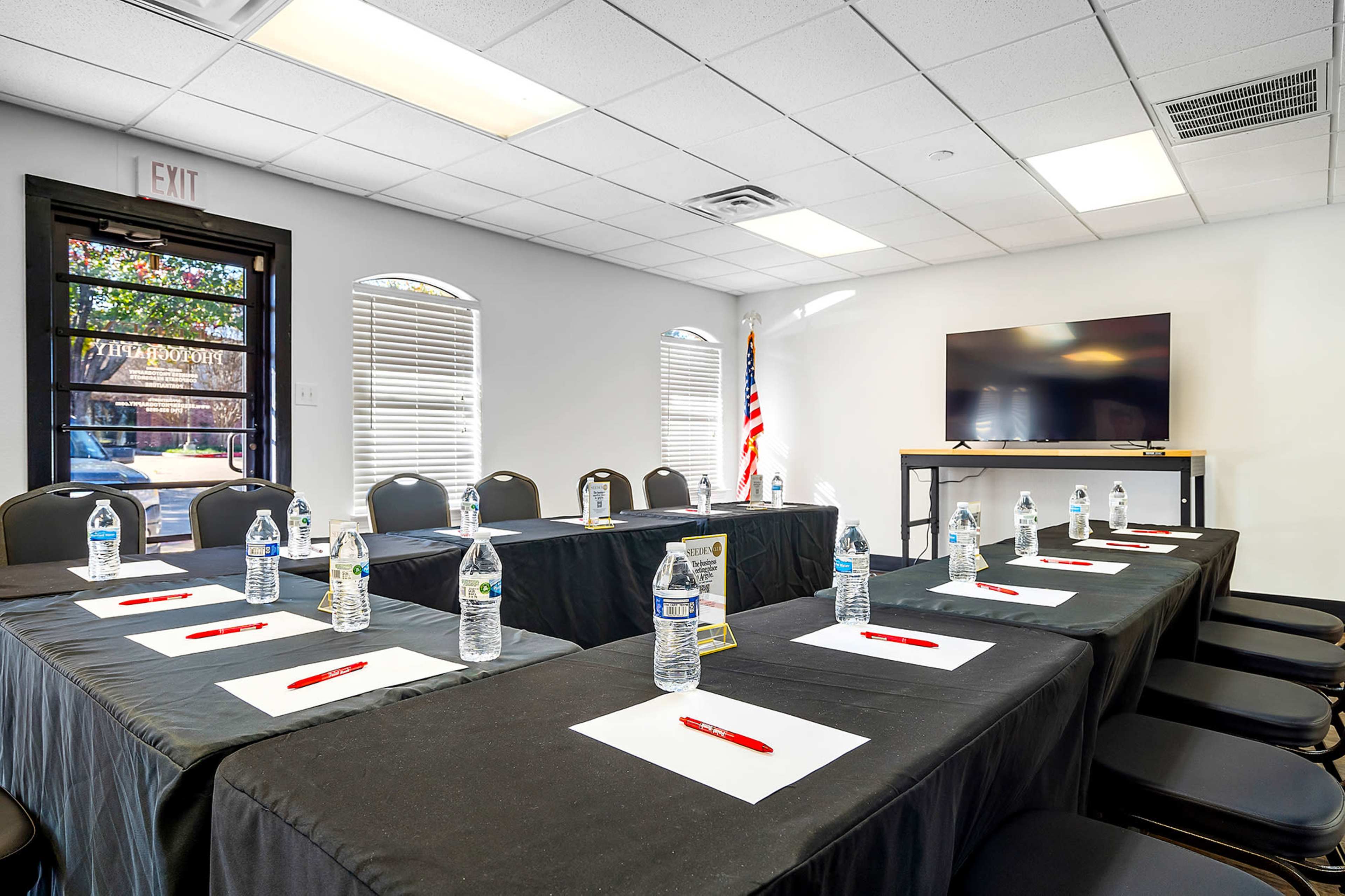A conference room is set up with black tables, chairs, water bottles, and red pens, facing a large screen and an American flag.