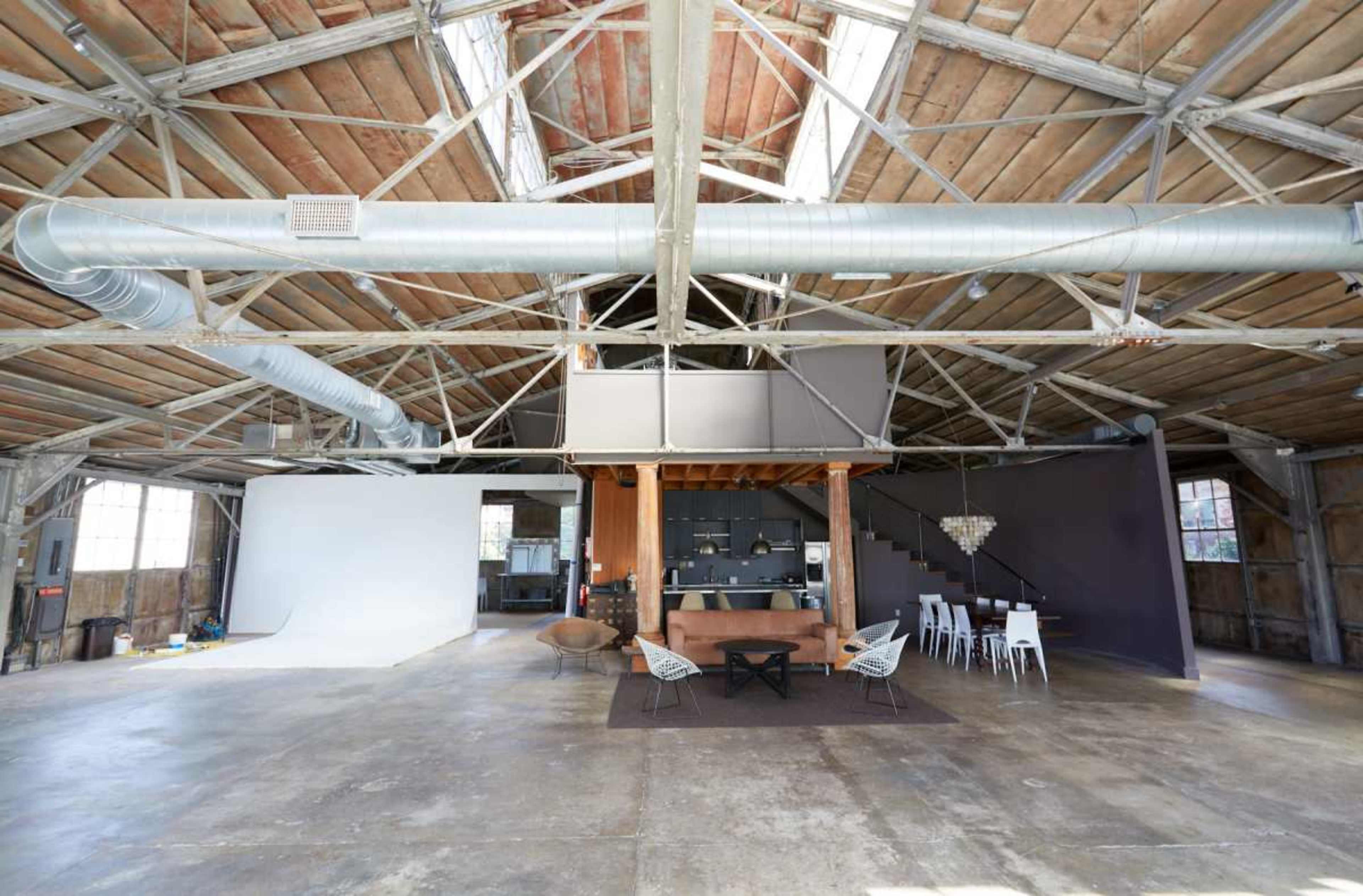 The image shows an open industrial-style space with high ceilings, large windows, and a mix of wood and metal materials, featuring a dining area and a kitchen.