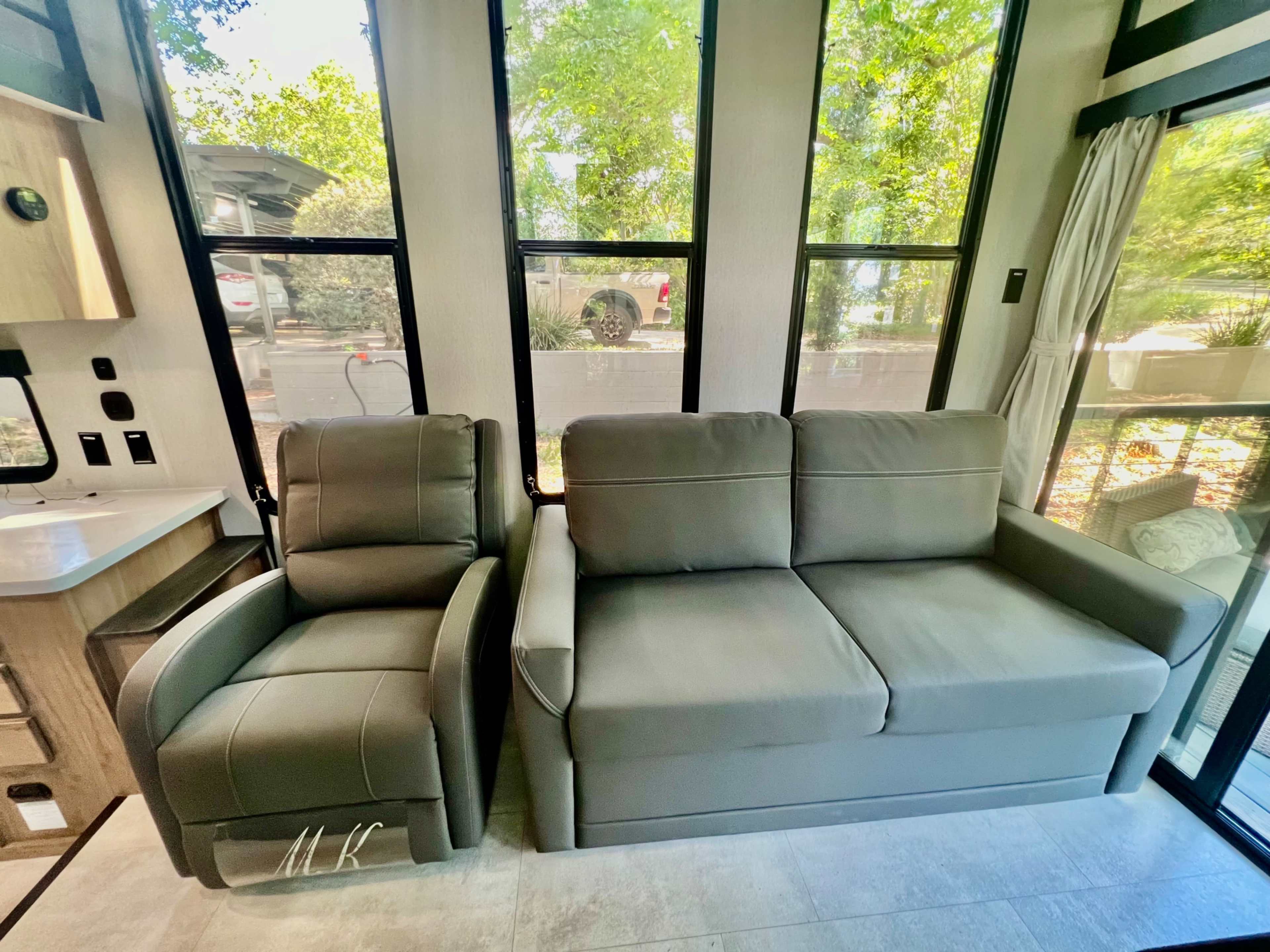 A modern living area featuring a gray couch and an armchair beside large windows that allow natural light to enter.