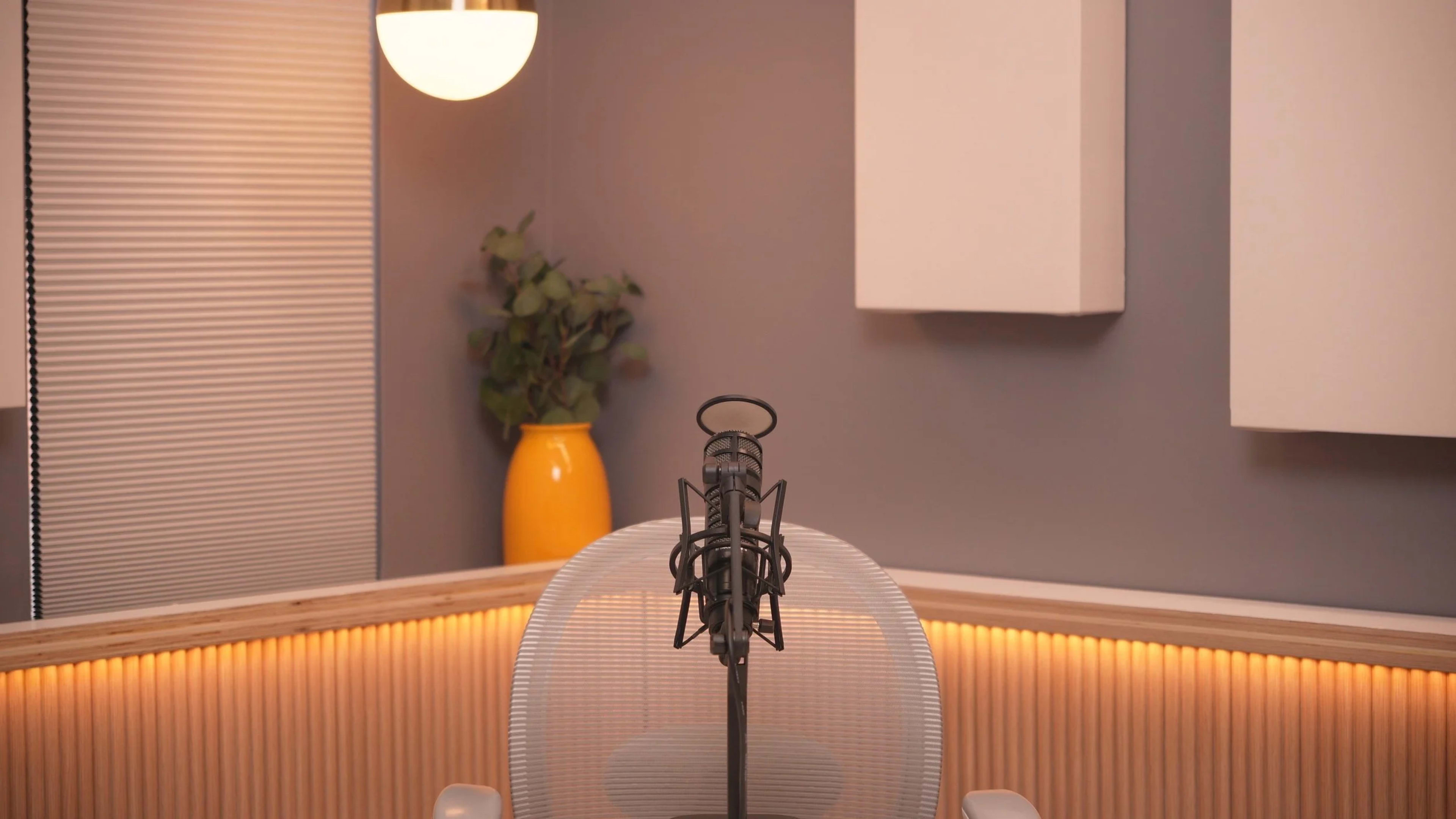 A microphone stands in front of a modern chair in a well-lit studio space featuring a plant and an orange vase against a gray wall.