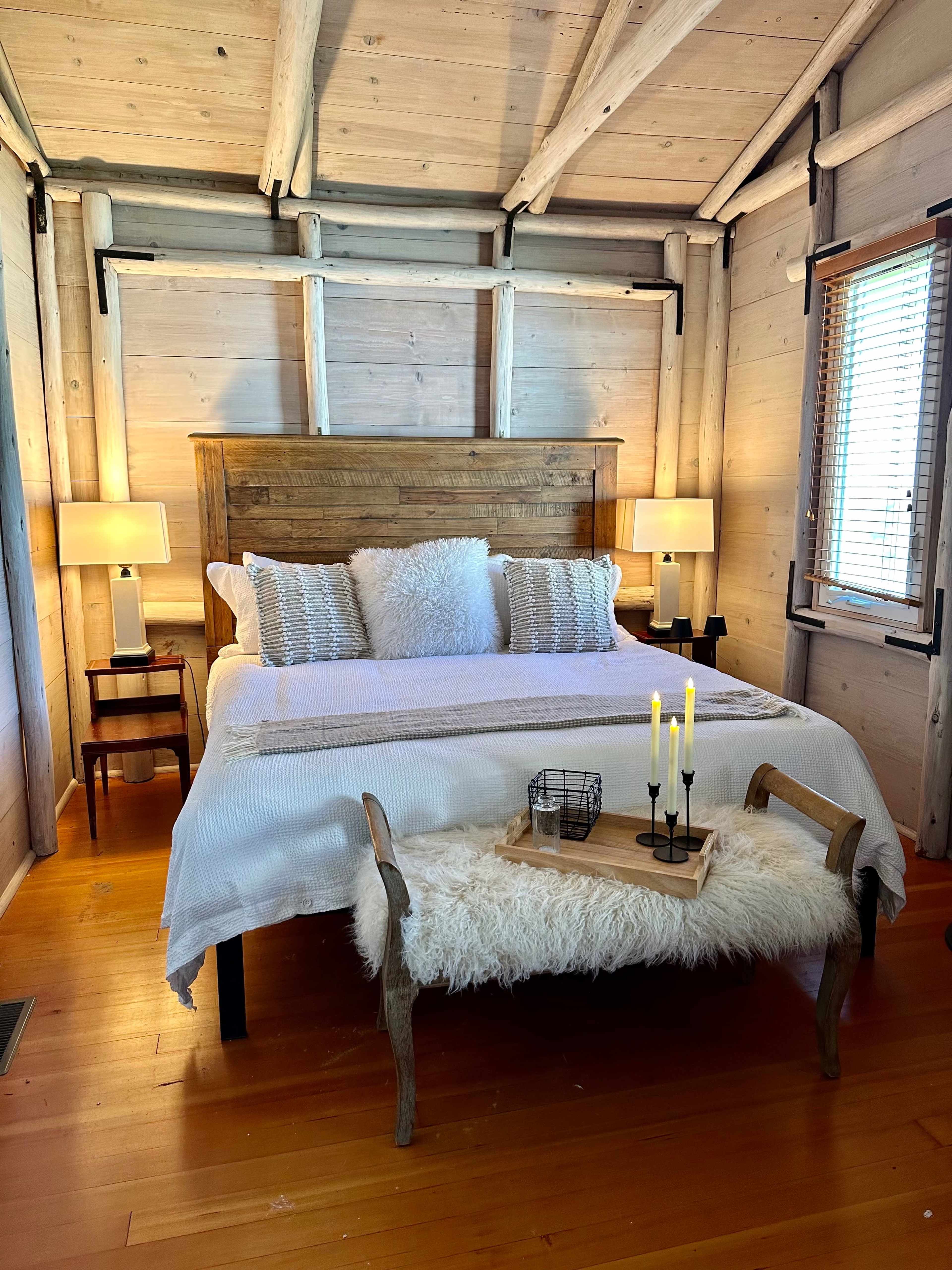 The bedroom features a wooden bed frame with white bedding, two bedside lamps, and a fur-covered bench at the foot of the bed.