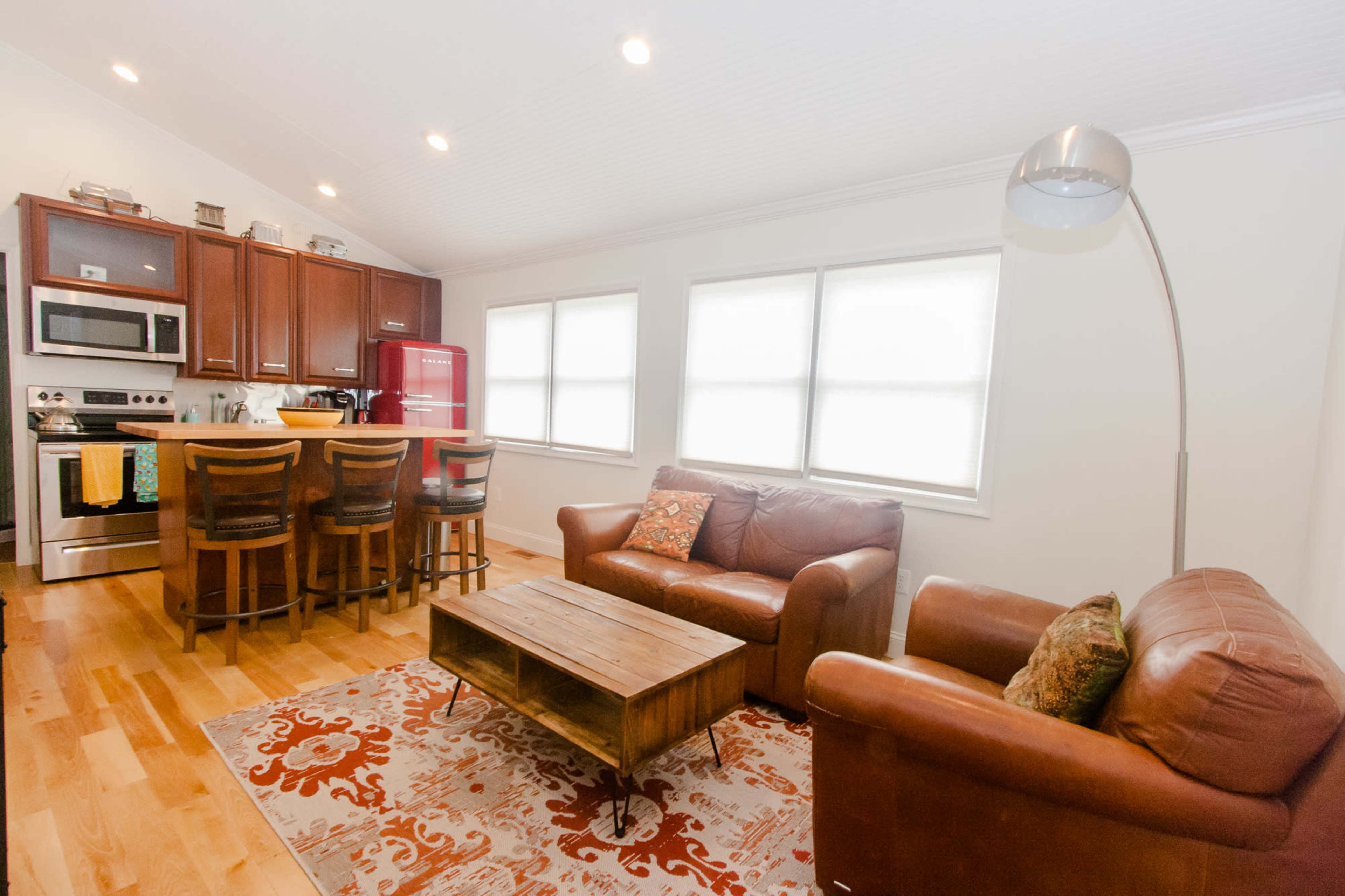 A cozy kitchen and living area with wooden flooring, featuring a red refrigerator, a seating area with brown leather sofas, and a wooden coffee table.
